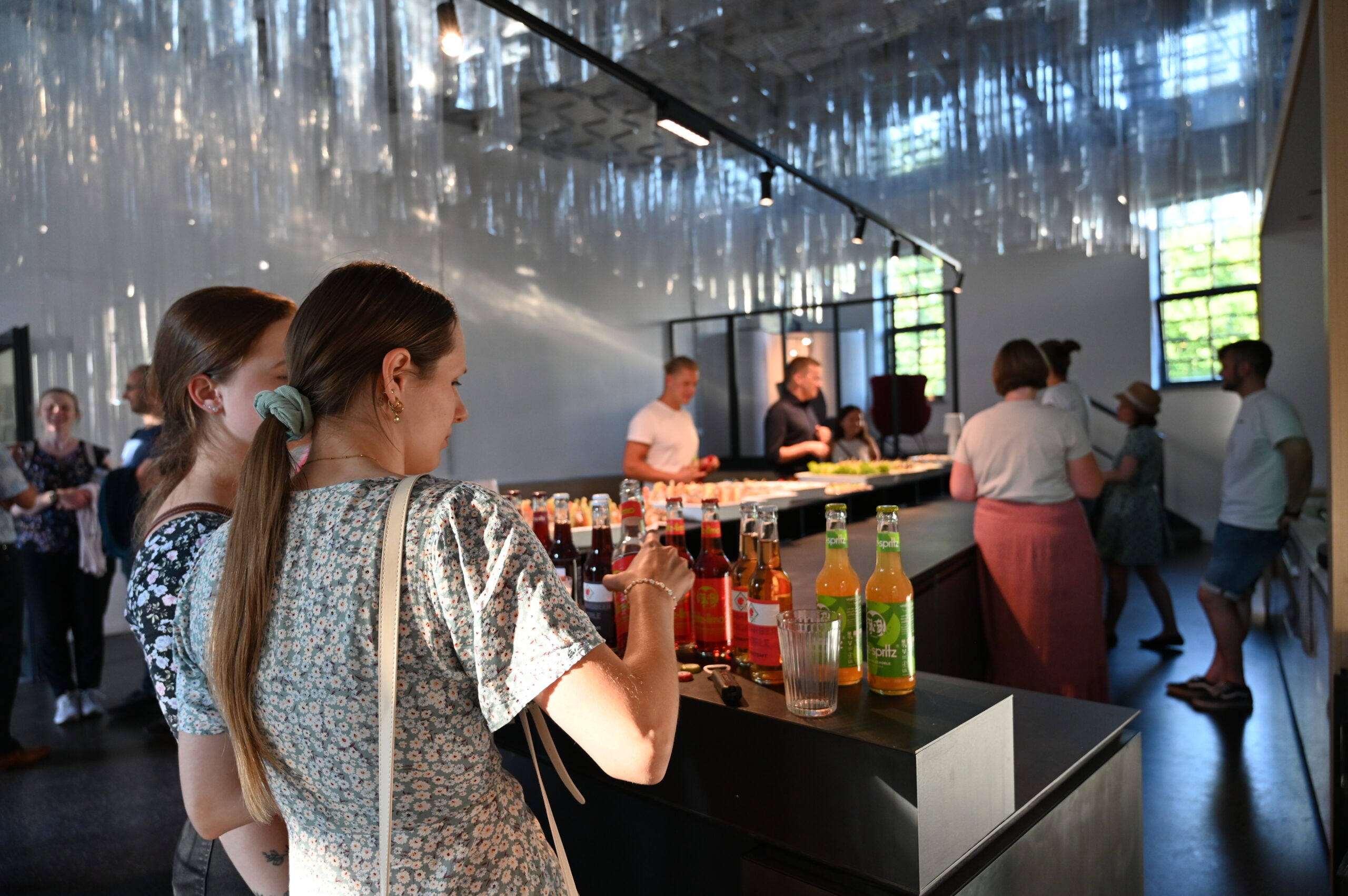 People are gathered in a modern bar at Hochschule Coburg, where various drinks are displayed on the counter. The room features sleek design elements with reflective materials on the ceiling, and sunlight illuminates the space through large windows.