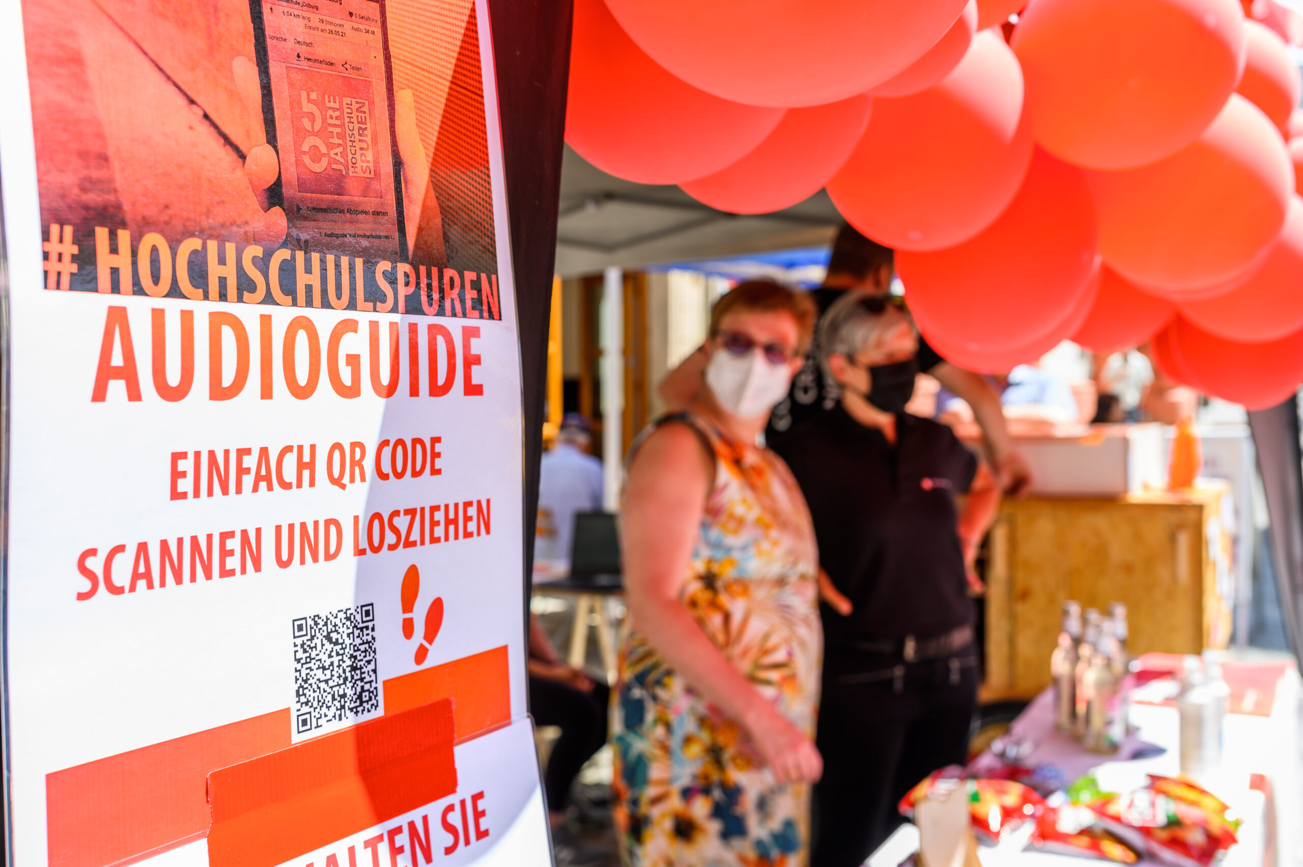 Ein Stand mit einem Schild, das für einen Audioguide per QR-Code wirbt, steht festlich geschmückt mit roten Luftballons. Zwei Menschen verweilen in der Nähe und entdecken den „#HOCHSCHULSPUREN AUDIOGUIDE“. Stolz mit der Hochschule Coburg verbunden, lädt er alle ein, einzigartige Geschichten und Einblicke zu entdecken.