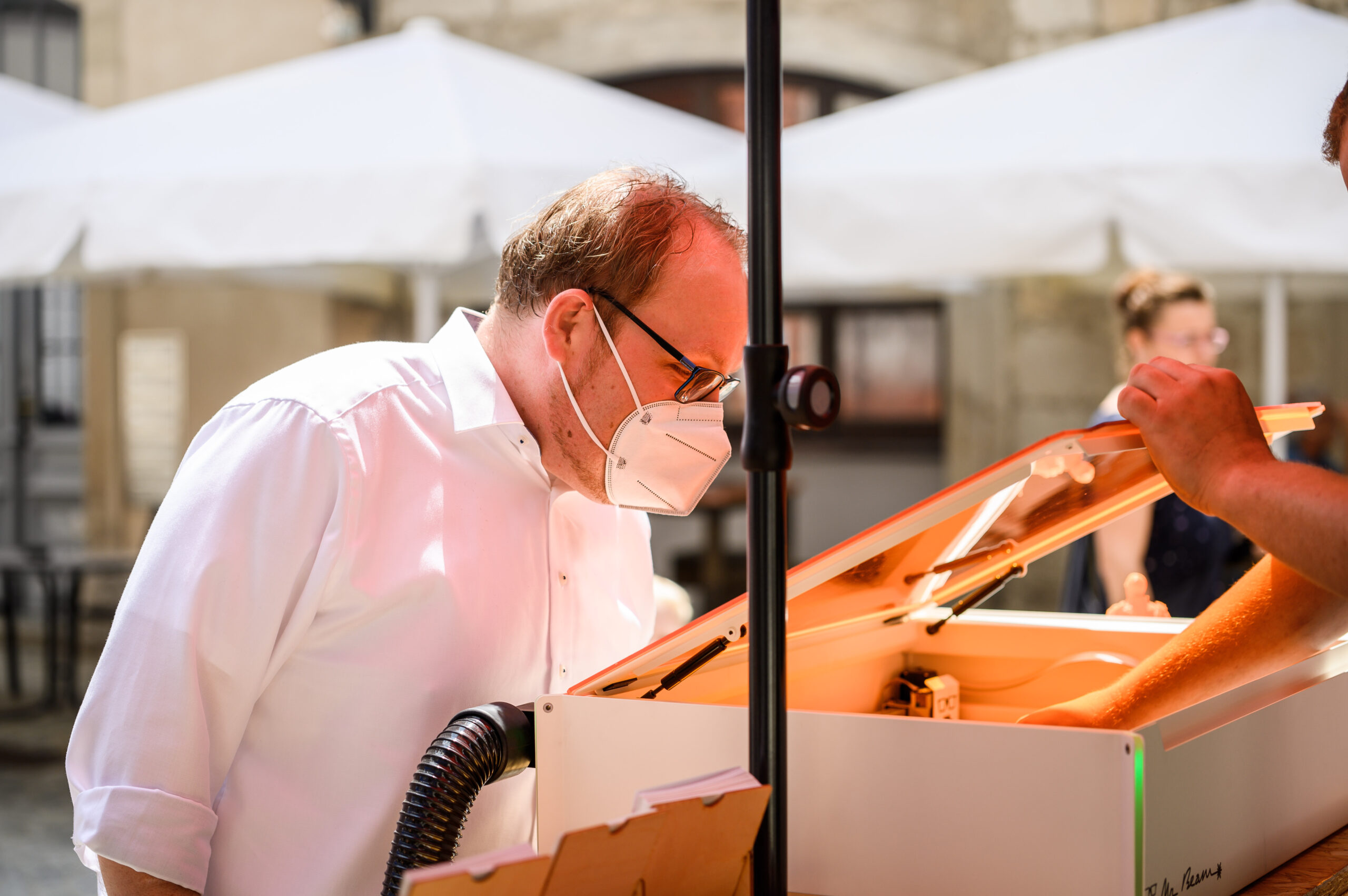 Ein Mann mit Maske und Brille, vermutlich von der Hochschule Coburg, inspiziert im Freien ein Gerät. Bei hellem Licht untersucht er den Inhalt genau, im Hintergrund sind Menschen und weiße Schirmdächer unscharf.