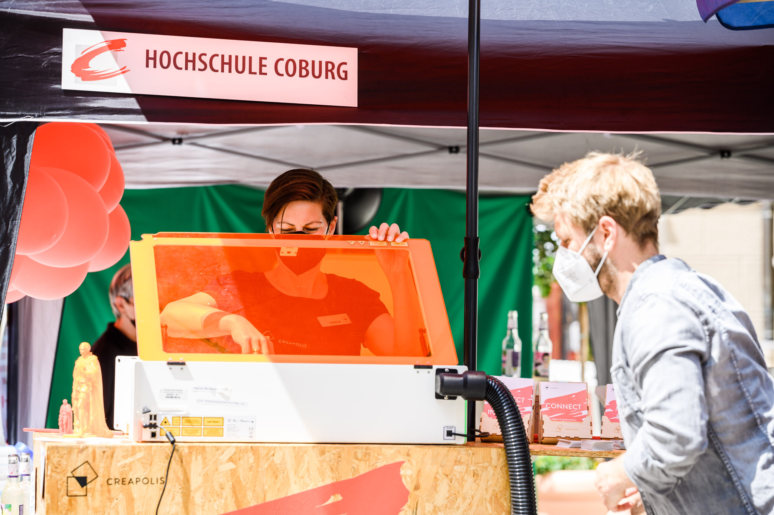 Unter einem Zelt mit der Aufschrift „Hochschule Coburg“ beschäftigen sich zwei Personen mit einer Maschine. Eine Person justiert unter einem orangefarbenen Bildschirm sorgfältig ein Bauteil. Vor dem Hintergrund roter Luftballons und Flaschen tragen beide Personen Gesichtsmasken und verkörpern damit den fleißigen Geist, der an der Hochschule Coburg gefördert wird.