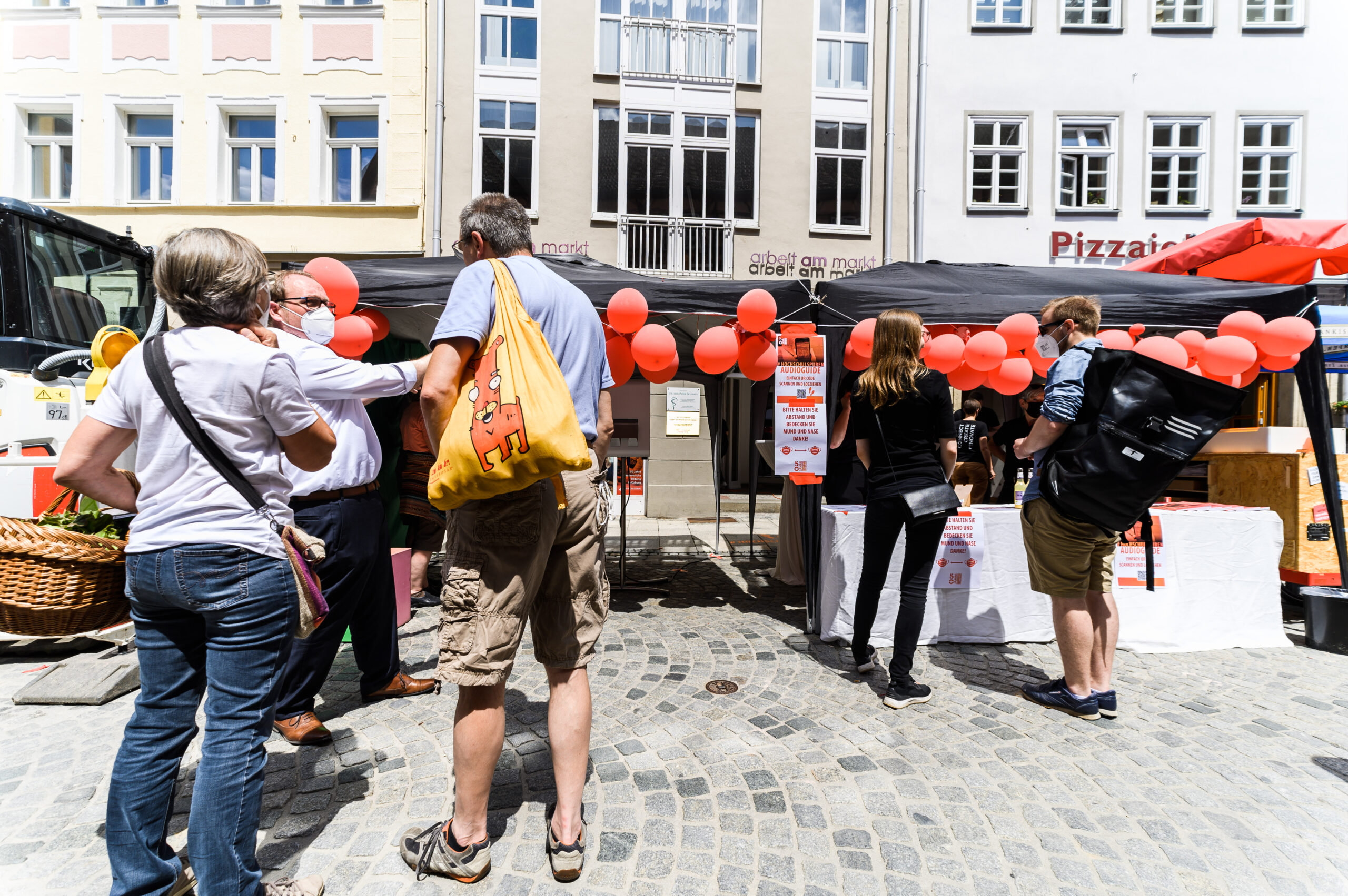 Menschen versammeln sich an einem Marktstand unter schwarzen, mit roten Luftballons geschmückten Vordächern, die an die Festtage an der Hochschule Coburg erinnern. In der Nähe stehen Körbe und eine Warenauslage, alles vor der Kulisse hoher Gebäude.