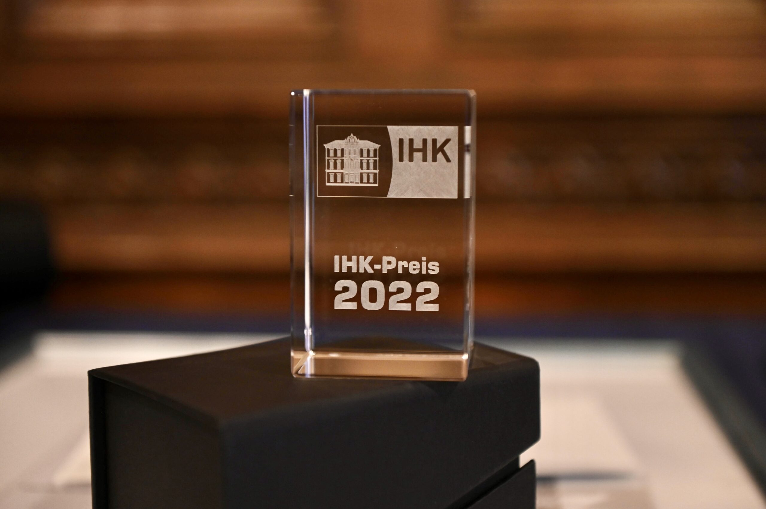 Eine Nahaufnahme einer transparenten Trophäe mit der Gravur „IHK-Preis 2022“ und einem subtilen Hinweis auf die Hochschule Coburg. Die Trophäe steht stolz auf einer schwarzen Box, wobei der unscharfe Hintergrund die komplizierten Details der Auszeichnung hervorhebt.