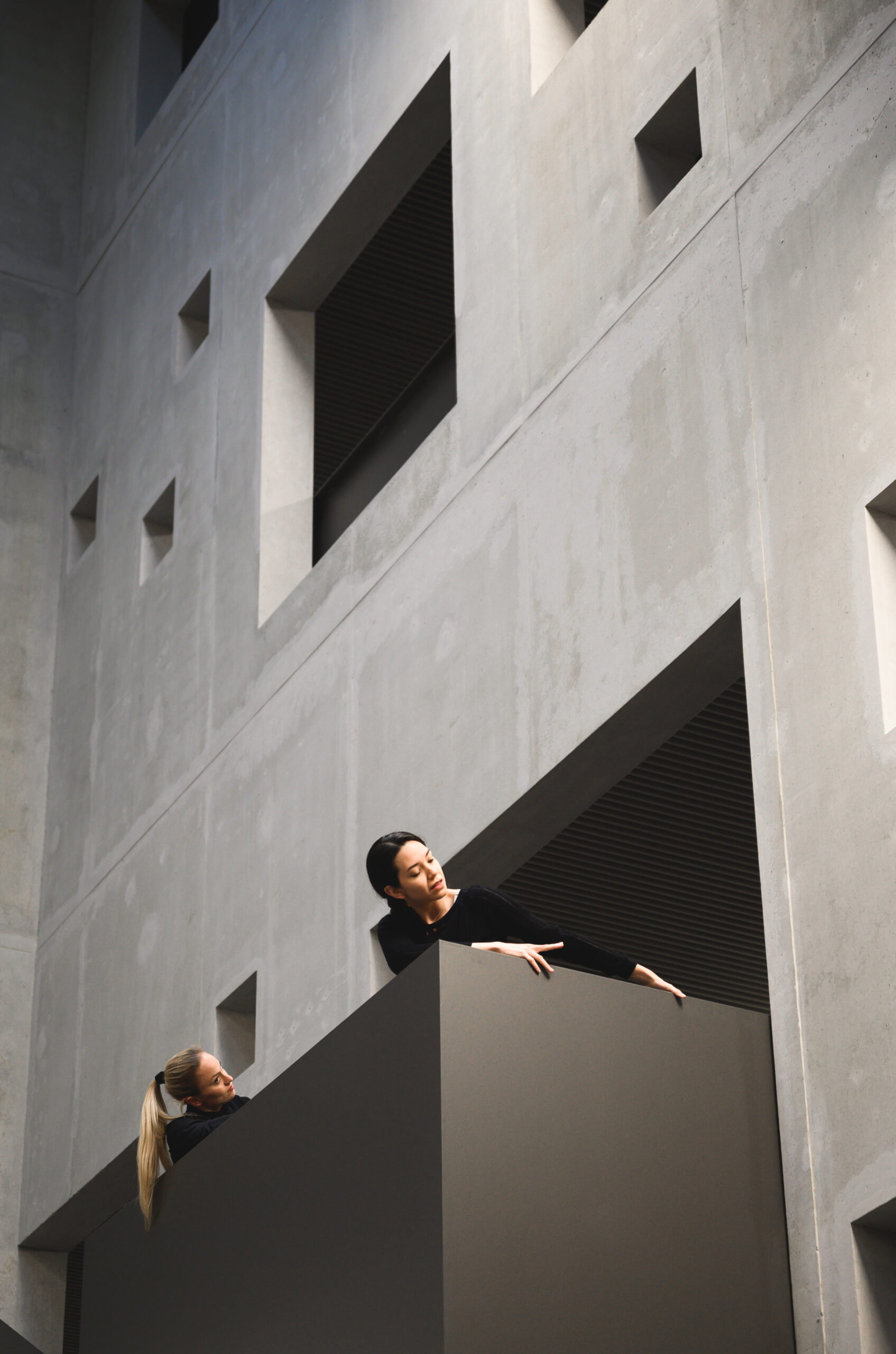 Hinter dem eckigen Betongebäude der Hochschule Coburg mit seinen zahlreichen quadratischen Fenstern interagieren zwei Menschen spielerisch miteinander. Das Design zeigt einen minimalistischen, modernen Architekturstil, wobei das Licht weiche Schatten auf die Oberflächen wirft.