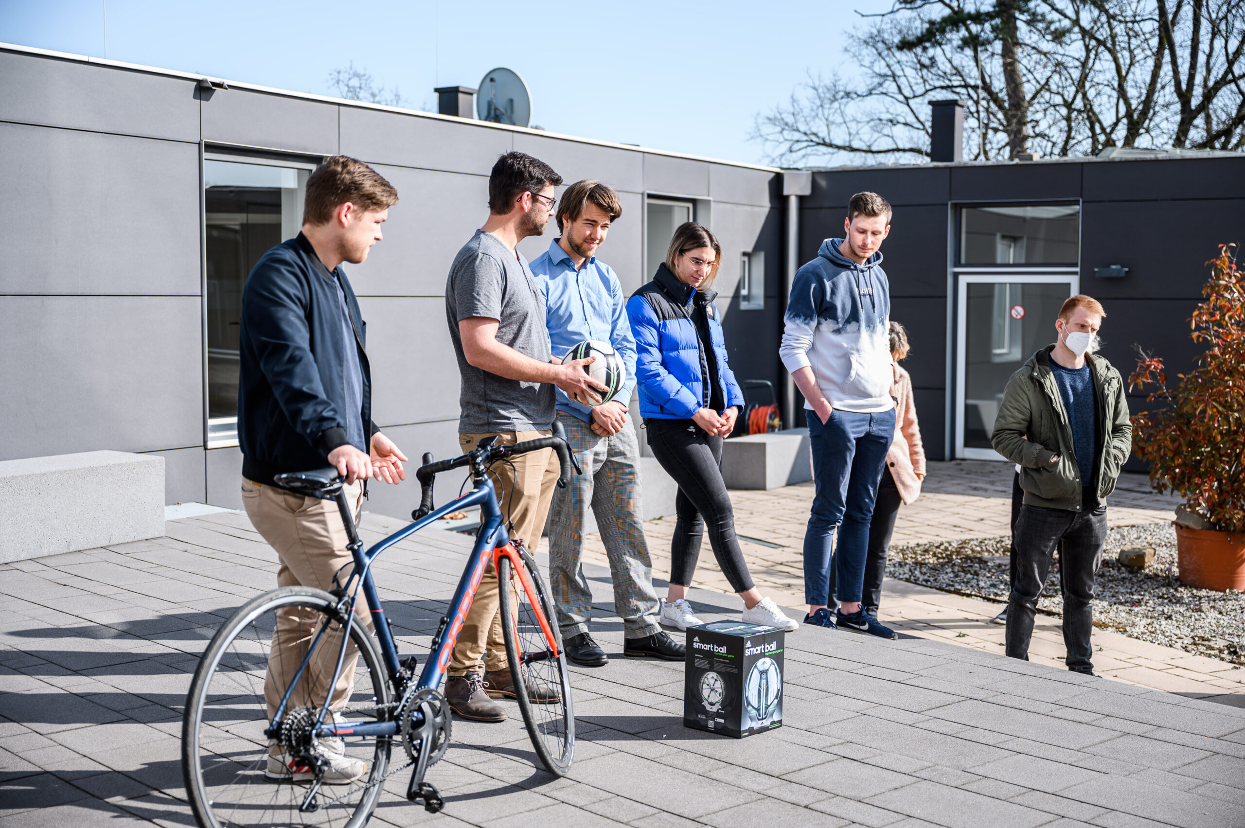 Eine Gruppe von Menschen versammelt sich draußen auf einer gepflasterten Fläche an der Hochschule Coburg. Eine Person hält ein Fahrrad neben einer schwarzen Box auf dem Boden. Vor der Kulisse eines modernen Gebäudes und eines klaren blauen Himmels scheinen sie in ein Gespräch vertieft zu sein.