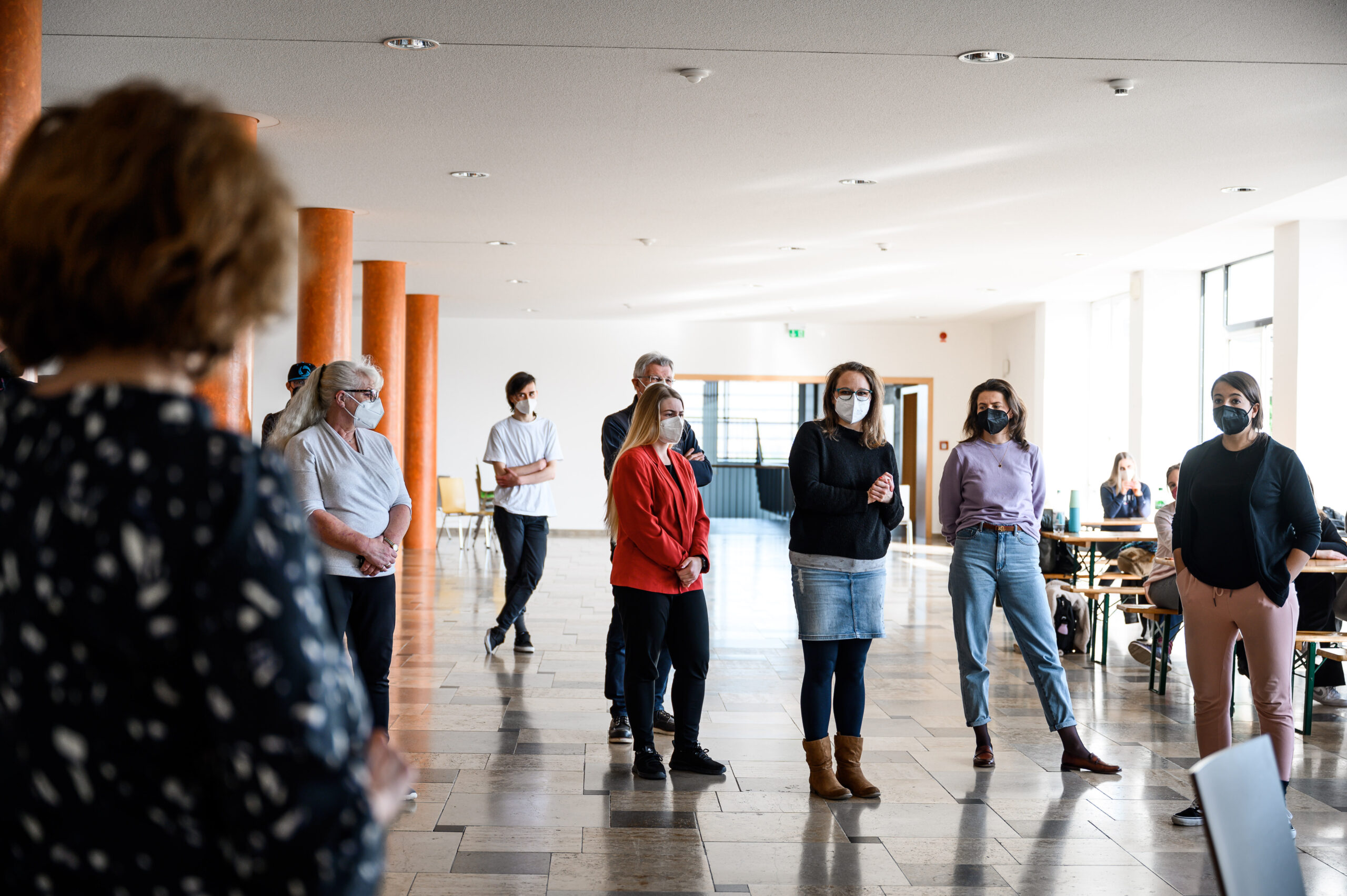 Eine Gruppe maskierter Menschen steht in einem großzügigen Raum mit großen Fenstern der Hochschule Coburg. Sie unterhalten sich, wobei eine Person die anderen anspricht. Im Hintergrund sind Tische und Stühle zu sehen, die den wissenschaftlichen Rahmen unterstreichen.