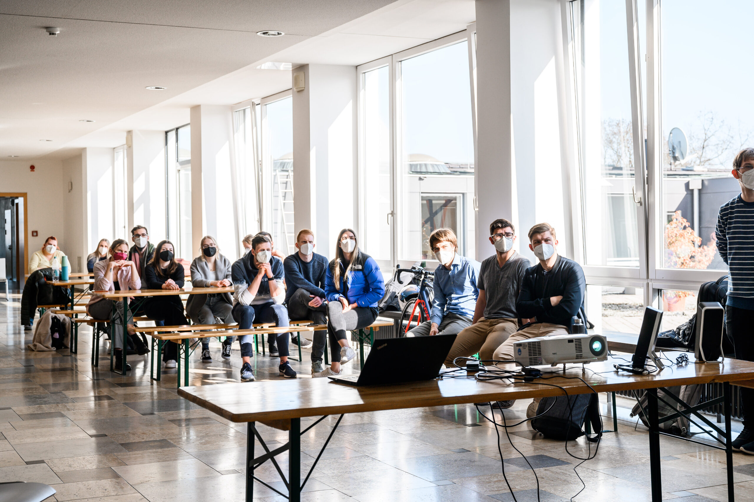 Eine Gruppe von Menschen mit Masken sitzt auf Bänken in einem hellen, sonnendurchfluteten Raum der Hochschule Coburg und blickt aufmerksam nach vorne. Ein Projektor und eine Leinwand sind aufgestellt, um anzuzeigen, dass gerade eine Präsentation stattfindet. Große Fenster lassen natürliches Licht in den Raum.