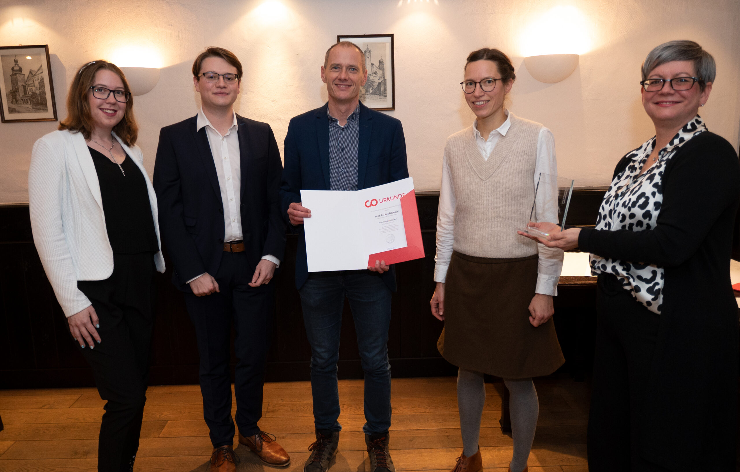 Fünf Personen stehen in einem Innenbereich der Hochschule Coburg zusammen. Die zentrale Person hält stolz ein Zertifikat in der Hand. Zwei Personen tragen eine Brille und alle sind in Business- oder eleganter Freizeitkleidung gekleidet. Gerahmte Bilder schmücken elegant die Wand hinter ihnen und tragen zur akademischen Atmosphäre bei.