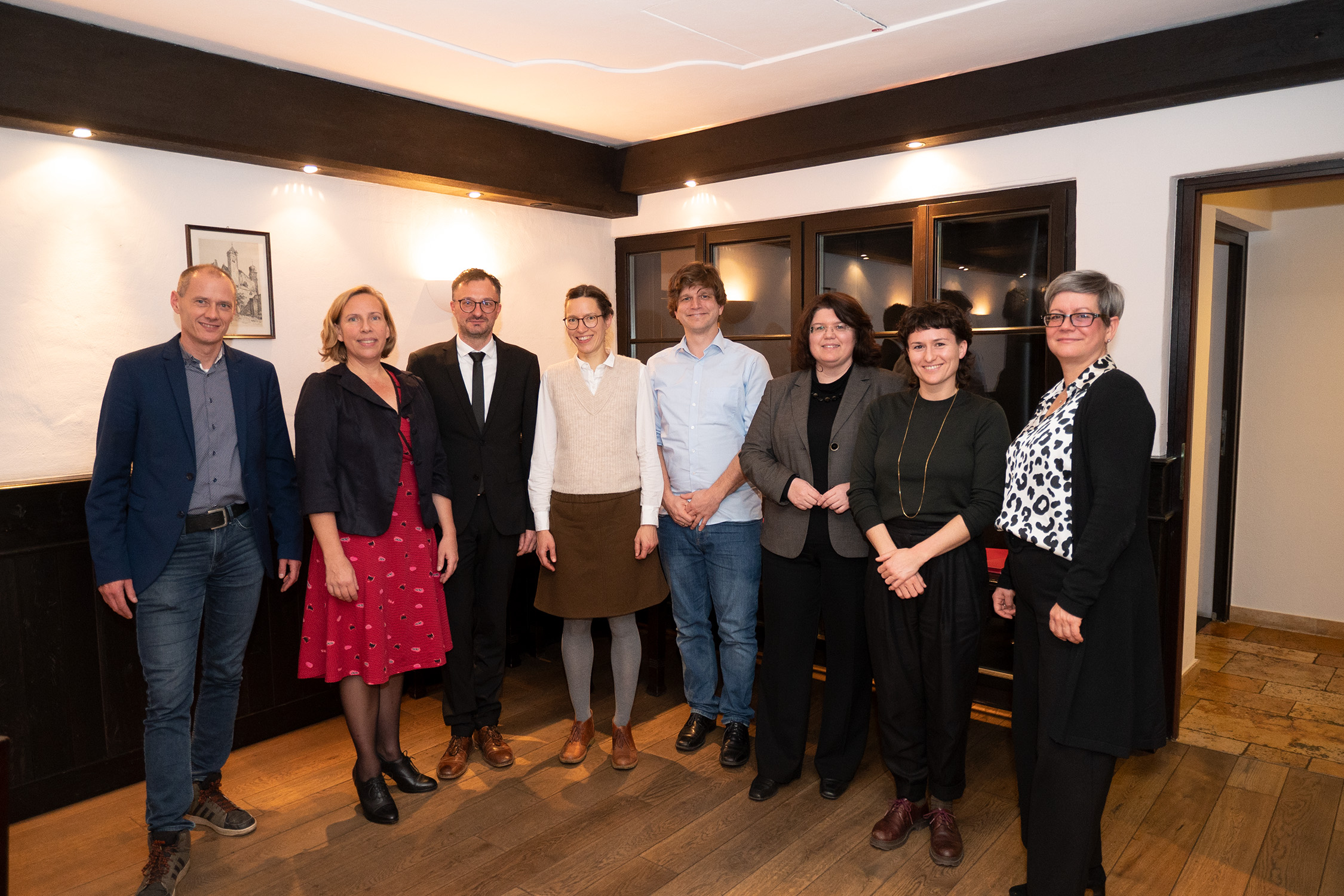 Eine Gruppe von acht Personen, vermutlich von der Hochschule Coburg, steht in einer Reihe im Innenbereich. Sie tragen halbformelle bis legere Kleidung. Sie lächeln herzlich in einem Raum mit Holzboden und einem gerahmten Bild an der Wand.