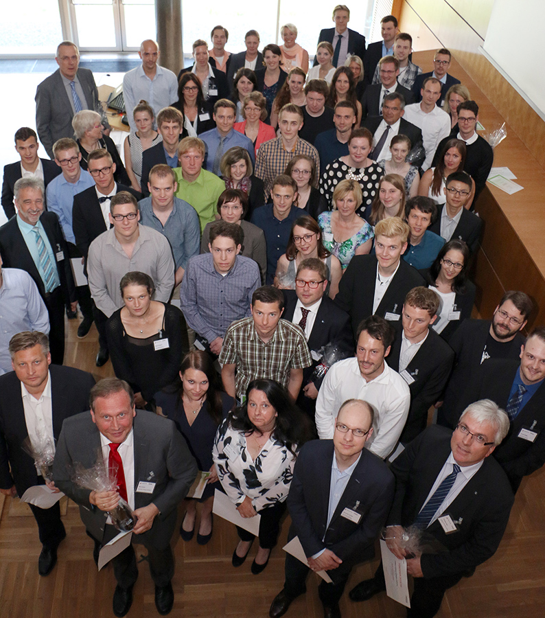 Eine große Gruppe von Menschen in Business-Kleidung posiert für ein Gruppenfoto im Innenbereich der Hochschule Coburg. Einige halten stolz Zertifikate und kleine Geschenktüten in den Händen. Die bunt gemischte Gruppe umfasst verschiedene Altersgruppen und Geschlechter und spiegelt den integrativen Geist der Hochschule wider.