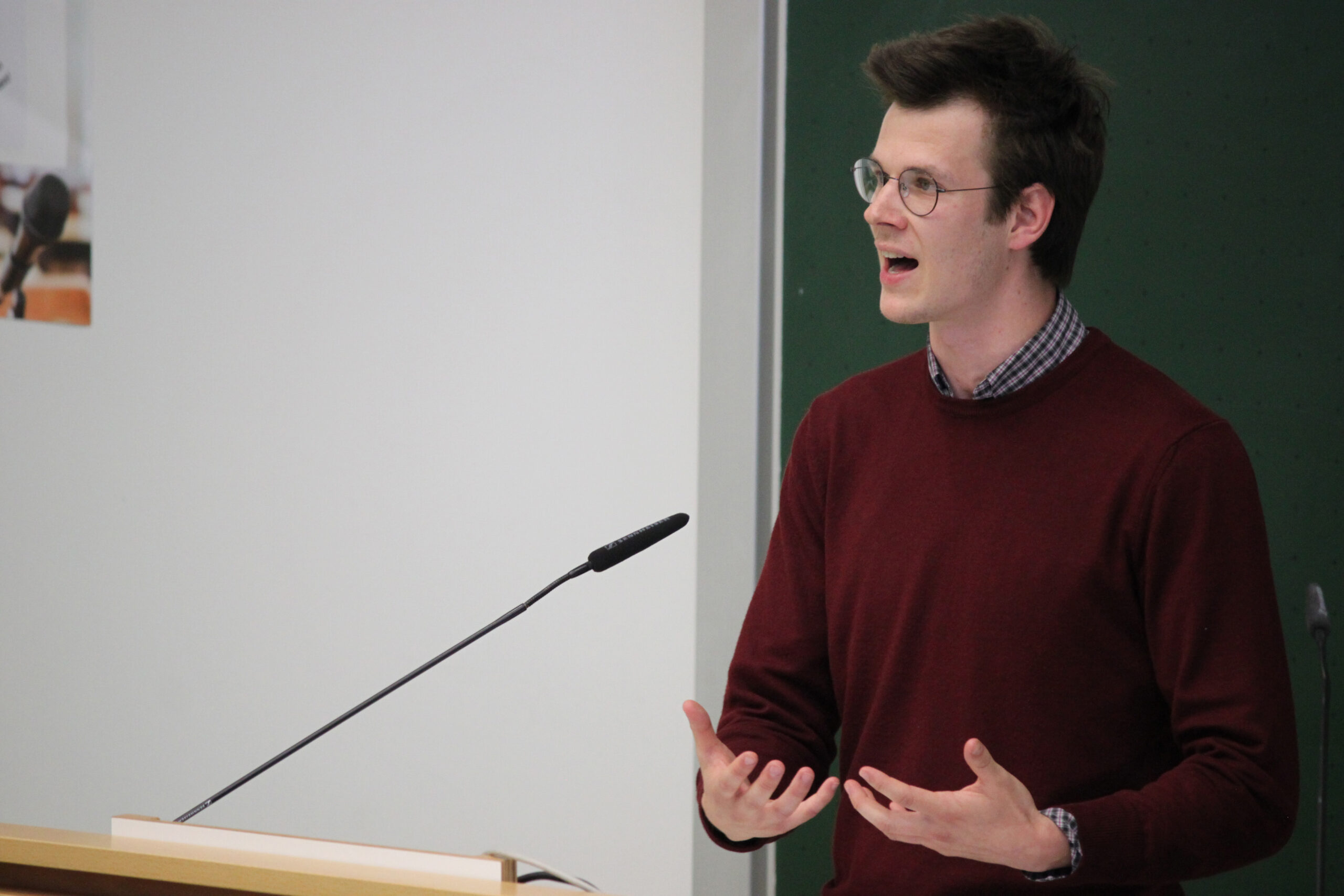 Eine Person mit Brille und kastanienbraunem Pullover spricht leidenschaftlich an einem Podium mit Mikrofon und gestikuliert, während sie sich an ein Publikum an der Hochschule Coburg wendet. Im Hintergrund ist eine grüne Tafel zu sehen.