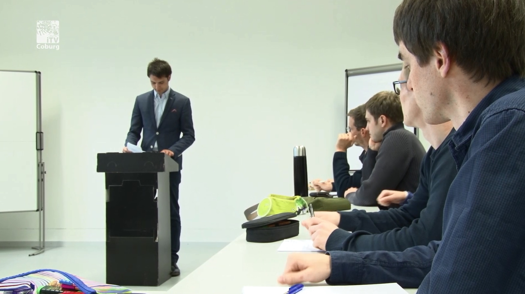 Ein Mann im Anzug steht an einem Podium vor mehreren sitzenden Personen, die sich Notizen machen, und verkörpert damit die akademische Atmosphäre der Hochschule Coburg. Der Raum mit seinen weißen Wänden und dem Whiteboard erinnert an ein Klassenzimmer oder eine Präsentationsumgebung. Verschiedene Gegenstände wie Stifte und Notizen liegen auf dem Tisch.