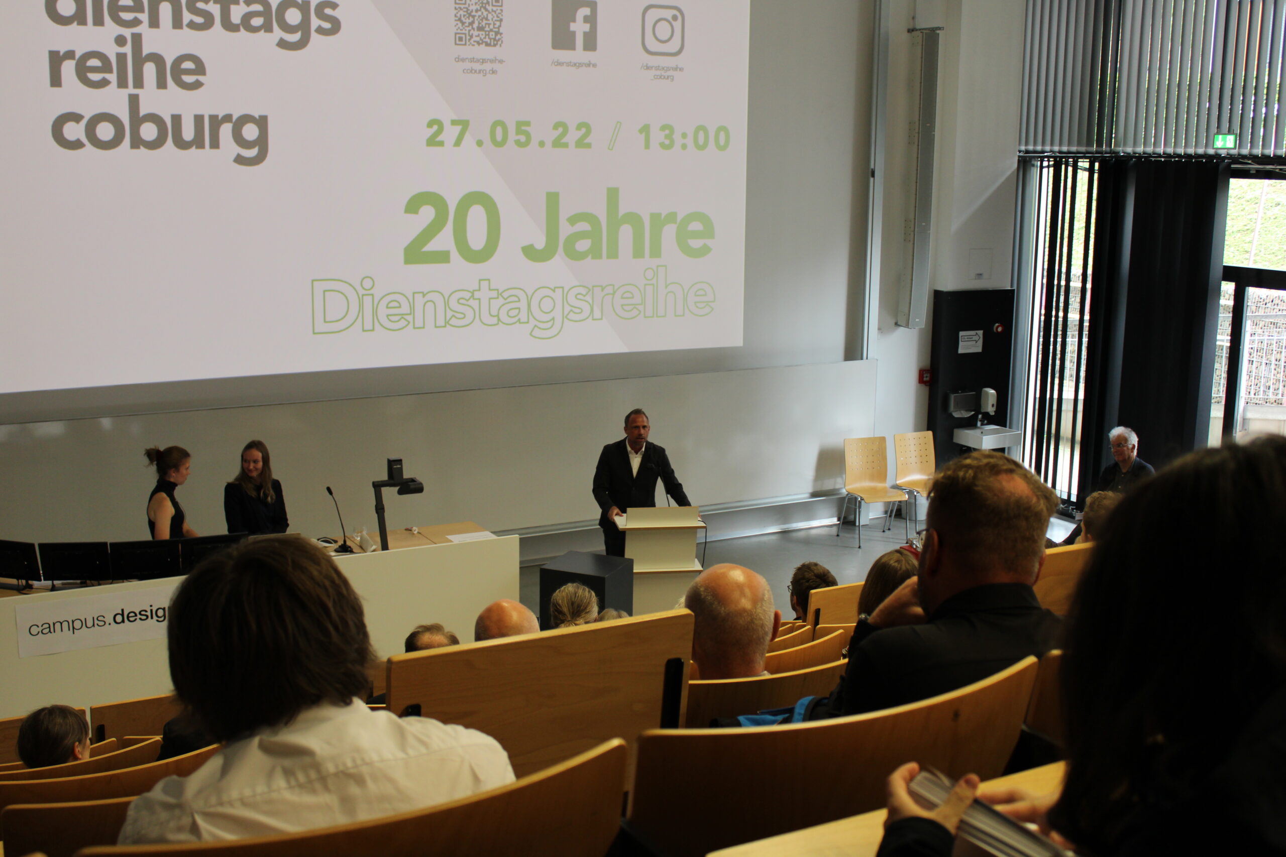 Ein Mann spricht an einem Podium in einem modernen Hörsaal der Hochschule Coburg während einer Veranstaltung zum Anlass von „20 Jahre Dienstagsreihe“. Die Teilnehmer sitzen, zwei Personen stehen daneben. Auf einem Bildschirm wird das Datum „27.05.22 / 13:00“ angezeigt, das auf den besonderen Anlass hinweist.