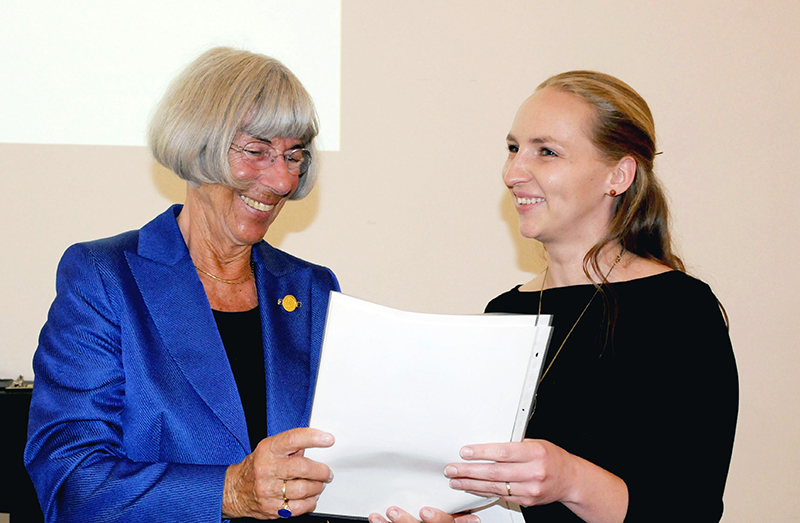 Zwei Frauen stehen nebeneinander und lächeln. Die ältere Frau links hat kurze graue Haare und trägt einen blauen Blazer, der die Hochschule Coburg repräsentiert. Die jüngere Frau rechts hat lange blonde Haare und trägt ein schwarzes Oberteil und hält ein Stück Papier in den Händen.