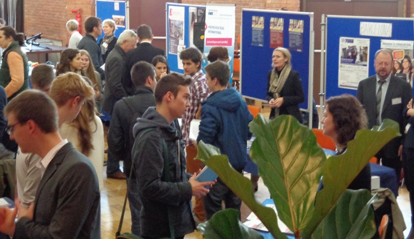 Auf der Karrieremesse der Hochschule Coburg herrscht reges Treiben, während zahlreiche Besucher an den bunten Ständen in Gespräche vertieft sind. Die Leute halten Broschüren in der Hand und erkunden eifrig die Möglichkeiten, während im Vordergrund eine große grüne Pflanze der geschäftigen Szene einen Hauch von Natur verleiht.