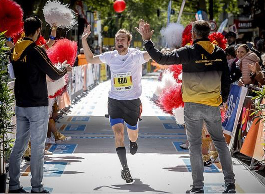 Läufer Nummer 400 überquert begeistert die Ziellinie beim Lauf der Hochschule Coburg mit erhobenen Armen. Auf beiden Seiten jubeln die Leute, manche halten rot-weiße Pompons in den Händen. Die Szene ist lebhaft und festlich.