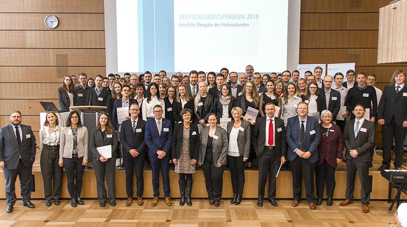 Eine große Gruppe von Menschen in formeller Kleidung steht in einem Konferenzraum der Hochschule Coburg zusammen. Im Hintergrund läuft auf einer Projektionsfläche das „Deutschlandstipendium 2018“. Viele halten Urkunden in den Händen, was auf eine offizielle Veranstaltung oder Preisverleihung schließen lässt.