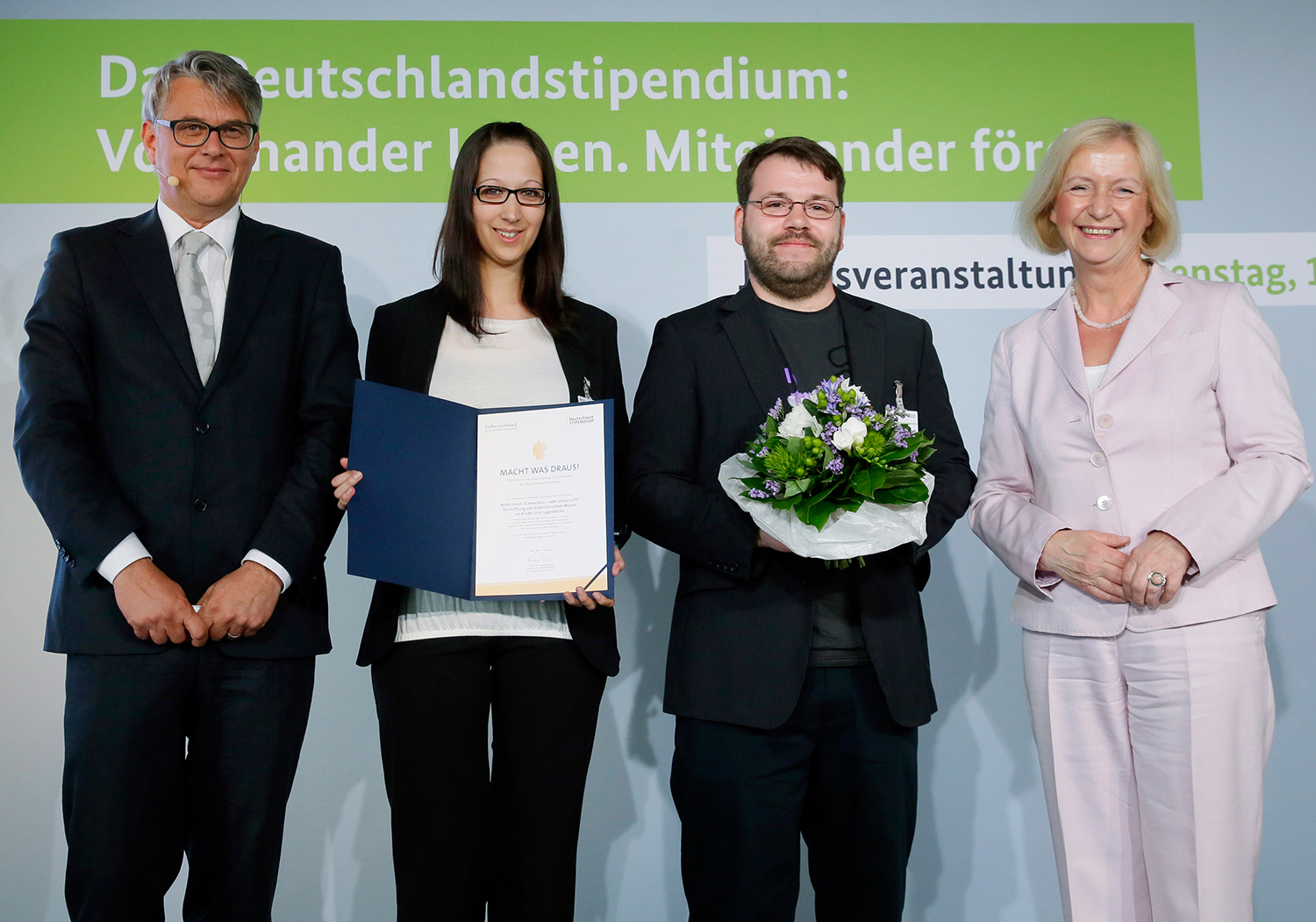 Vier Personen stehen bei einer Preisverleihung vor einem Banner der Hochschule Coburg. Eine Frau hält eine Urkunde in der Hand, ein Mann überreicht ihr einen Blumenstrauß. Alle lächeln und sind in formeller Kleidung gekleidet.