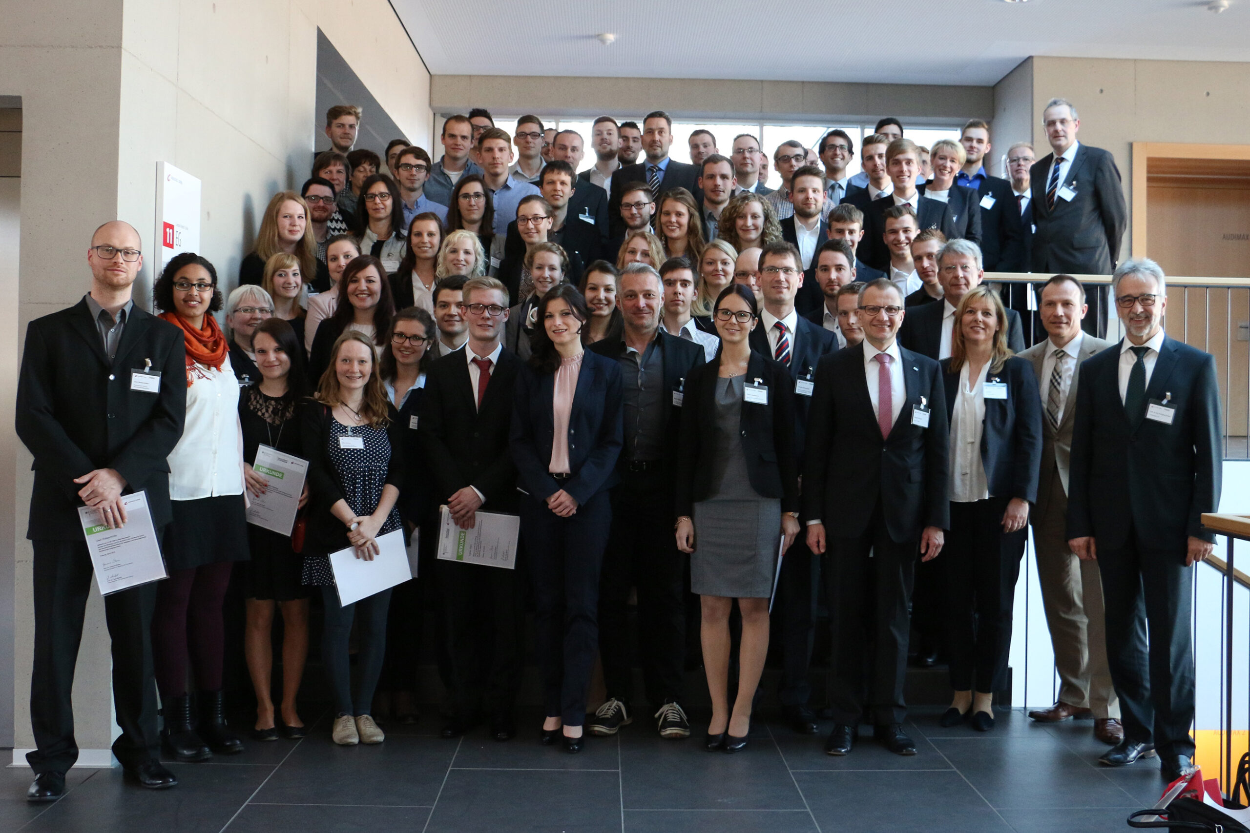 Eine große Gruppe professionell gekleideter Menschen posiert für ein Gruppenfoto auf der Treppe der Hochschule Coburg. Einige halten Papiere in den Händen, einige tragen Namensschilder. Der formelle Rahmen lässt vermuten, dass es sich um eine Konferenz oder ein Seminar handeln könnte.