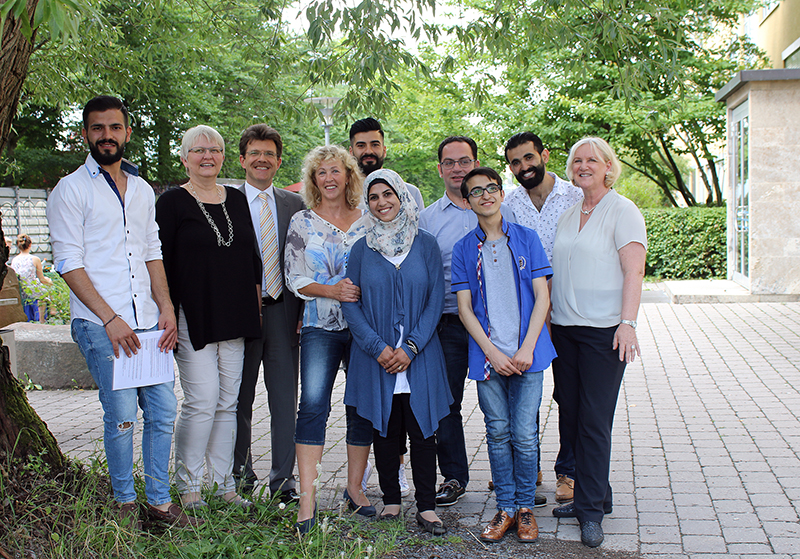 Eine Gruppe von zehn Personen steht im Freien auf einem gepflasterten Weg, umgeben von Grün. Sie repräsentieren die Hochschule Coburg. Sie posieren für ein Foto, lächeln und scheinen unterschiedlicher Herkunft und unterschiedlichen Alters zu sein. Sie tragen Freizeit- und Businesskleidung.