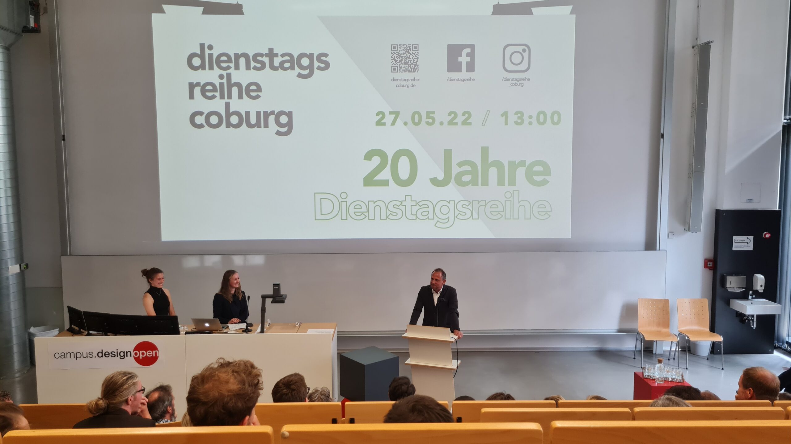 In einem modernen Hörsaal der Hochschule Coburg findet ein Vortrag statt, auf einer großen Leinwand werden Informationen zur Diensttagsreihe Coburg und ihrer 20-jährigen Jubiläumsveranstaltung am 27.05.22 um 13:00 Uhr angezeigt. Das Publikum sitzt und hört aufmerksam zu.