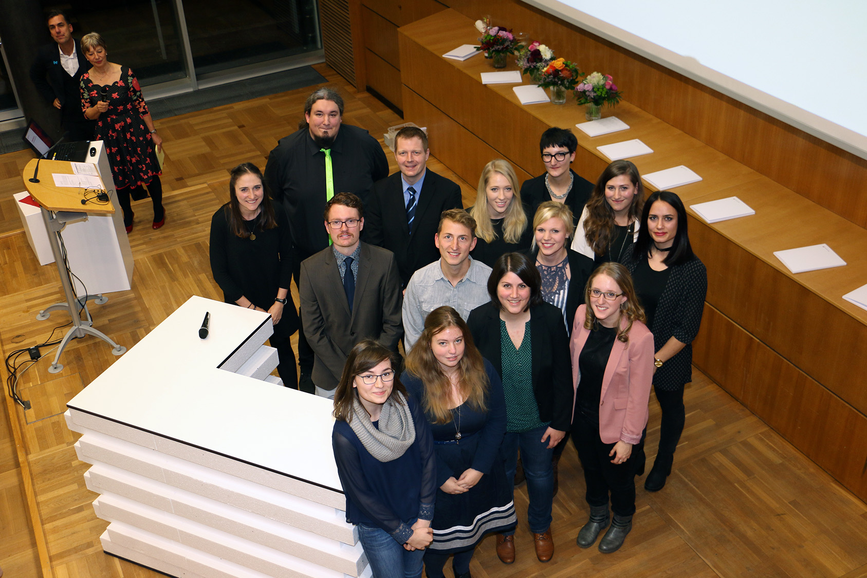 Eine Gruppe der Hochschule Coburg steht in einem Konferenzraum zusammen, in Reihen auf einem Holzboden in der Nähe des Podiums angeordnet. Einige Personen tragen formelle Kleidung, was zur formellen Atmosphäre beiträgt. Eine Reihe von Blumen und Papieren schmückt einen Sims hinter ihnen.