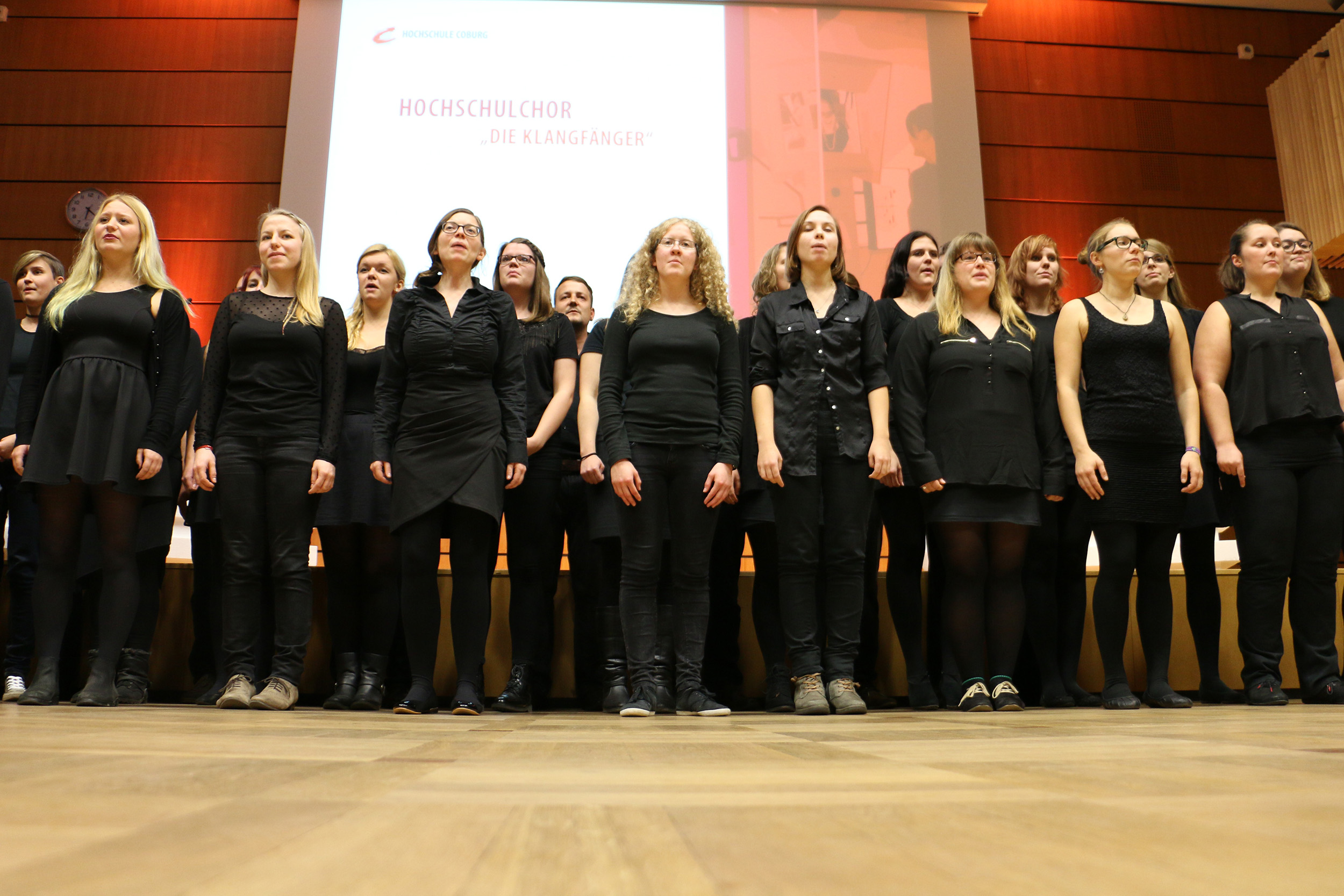 Ein Chor aus etwa zwölf Frauen in schwarzen Kostümen steht vor einer Leinwand mit der Aufschrift „Hochschulchor ‚Die Klangfänger‘“, der offenbar der Hochschule Coburg angeschlossen ist. Sie sind konzentriert und treten in einem warmen, holzgetäfelten Raum auf.