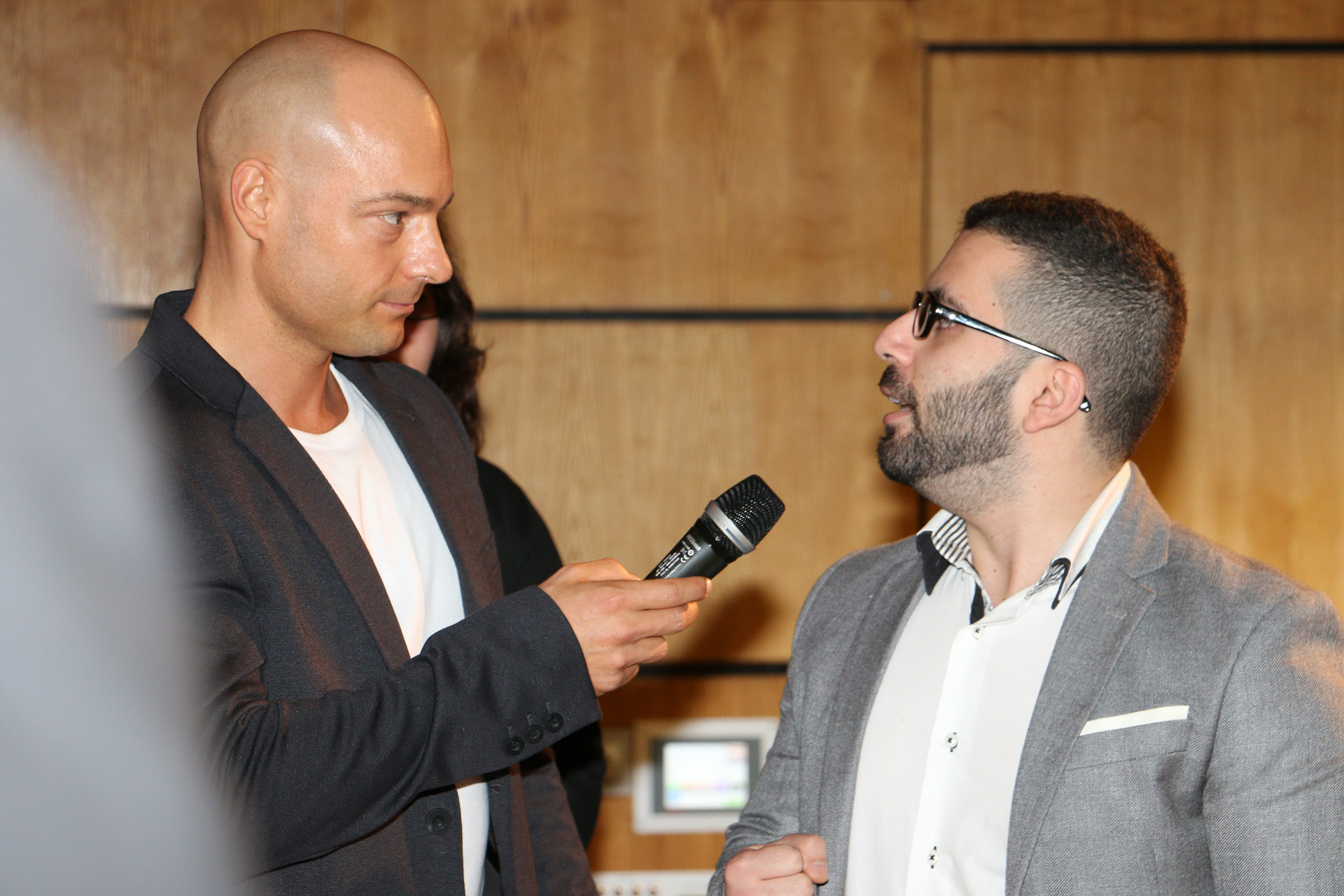 Ein kahlköpfiger Mann in einem schwarzen Anzug hält ein Mikrofon und interviewt einen Mann mit Brille und Bart, der einen grauen Blazer trägt. Sie befinden sich in einem Raum mit Holzwänden, möglicherweise an der Hochschule Coburg.