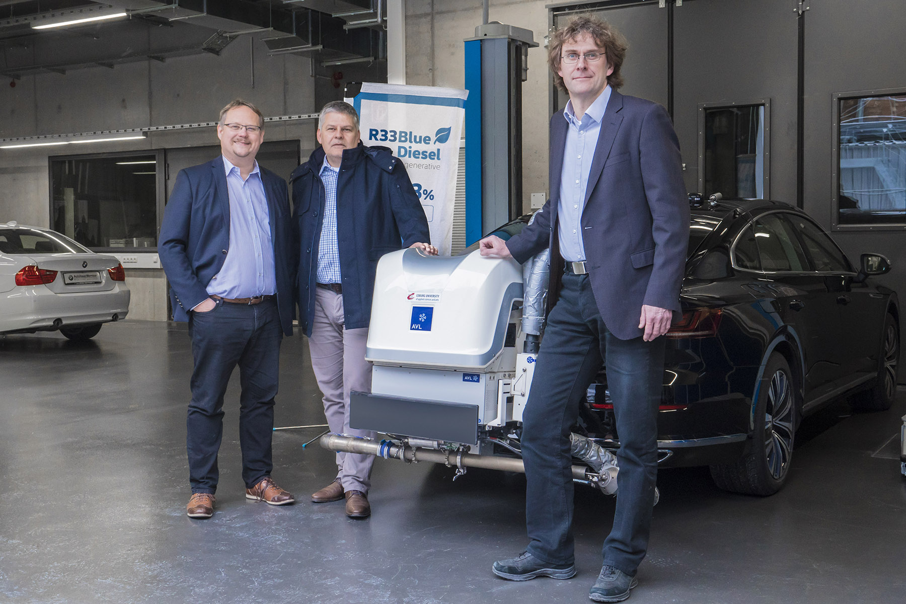 Drei Männer in Business-Kleidung stehen in einer Garage neben einem schwarzen Auto mit der Aufschrift „R33 Blue Diesel“. Im Hintergrund sind ein weiteres Auto und Industrieanlagen zu sehen, was den Innovationsgeist der Hochschule Coburg widerspiegelt.