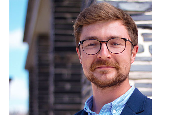 Ein Mann mit kurzen braunen Haaren, Brille und Bart lächelt leicht. Er trägt einen blauen Blazer und ein hellblaues Hemd. Hinter ihm ragt das verschwommene Backsteingebäude der Hochschule Coburg unter einem teilweise bewölkten Himmel empor.