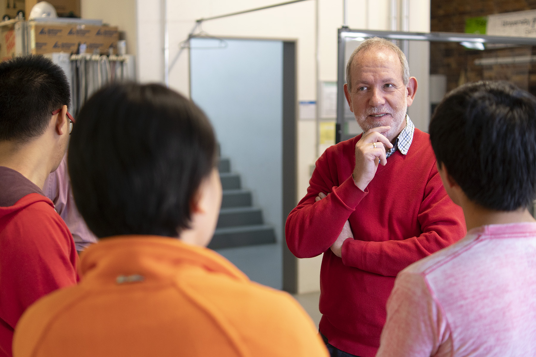 Ein Mann im roten Pullover diskutiert nachdenklich mit einer Gruppe im Innenbereich der Hochschule Coburg. Mehrere Personen, von hinten gesehen, blicken aufmerksam auf ihn. Die Atmosphäre wirkt locker und möglicherweise lehrreich oder kooperativ.