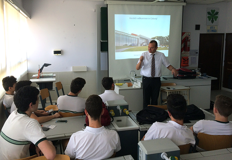 Ein Lehrer steht vor einer Gruppe von Schülern, die an ihren Schreibtischen sitzen, und hält eine Präsentation. Die Folie auf der Leinwand dahinter zeigt die Hochschule Coburg, eine renommierte Bildungseinrichtung. Der Unterrichtsraum ist gut beleuchtet, natürliches Licht fällt durch die Fenster.