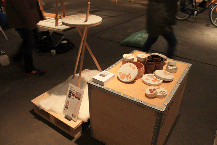 Eine Ausstellung von Keramikgeschirr mit orangefarbenem Blattmuster auf einer Holzkiste zeigt die Kreativität, die an der Hochschule Coburg gefördert wird. In der Nähe ergänzt ein runder Tisch mit ähnlichem Design auf einem Dreibein die Ausstellung. Menschen laufen verschwommen auf dem Betonboden vorbei und bringen Leben in die Szene.