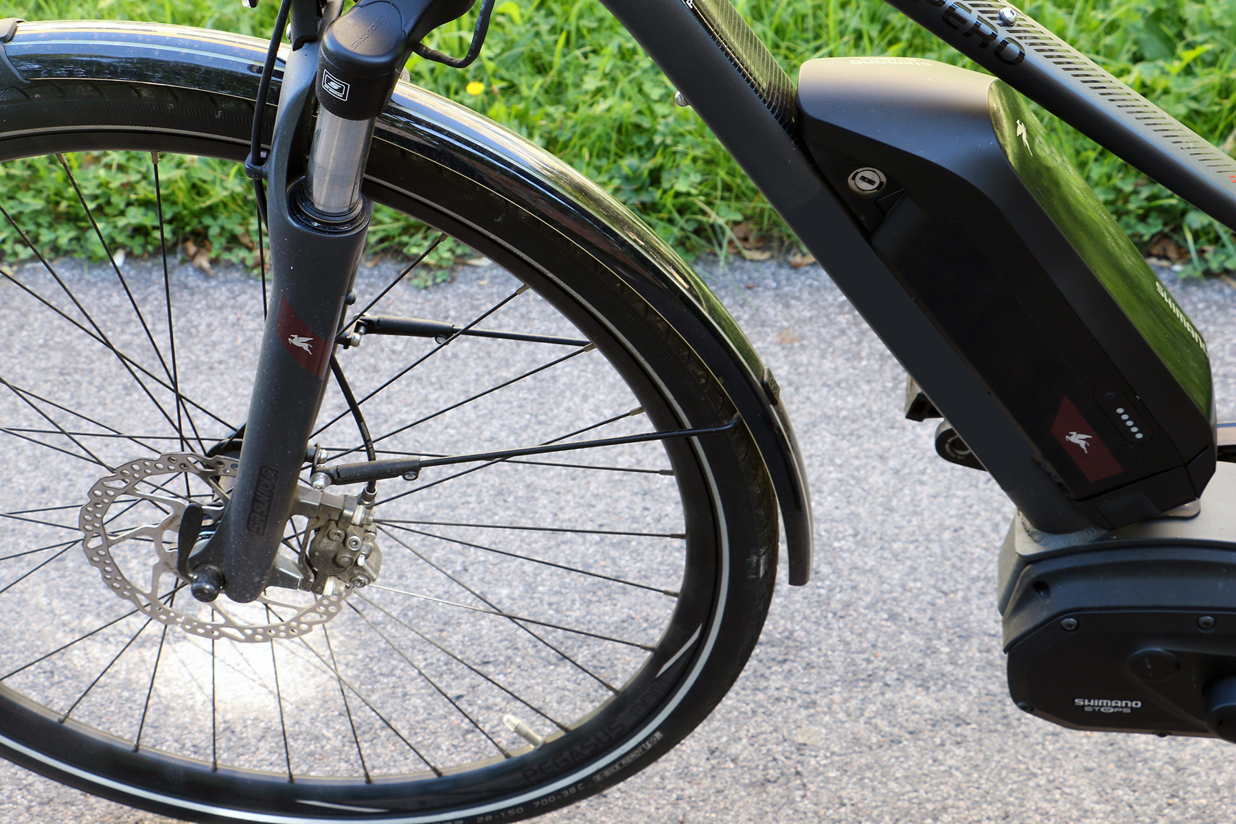 Nahaufnahme des Vorderrads und des Rahmens eines Elektrofahrrads in der Nähe der Hochschule Coburg. Der Akku des Fahrrads ist am schwarzen Rahmen über den Pedalen montiert. Am Rad sind Scheibenbremsen zu sehen, im Hintergrund ist ein gepflasterter Weg mit Gras daneben zu sehen.