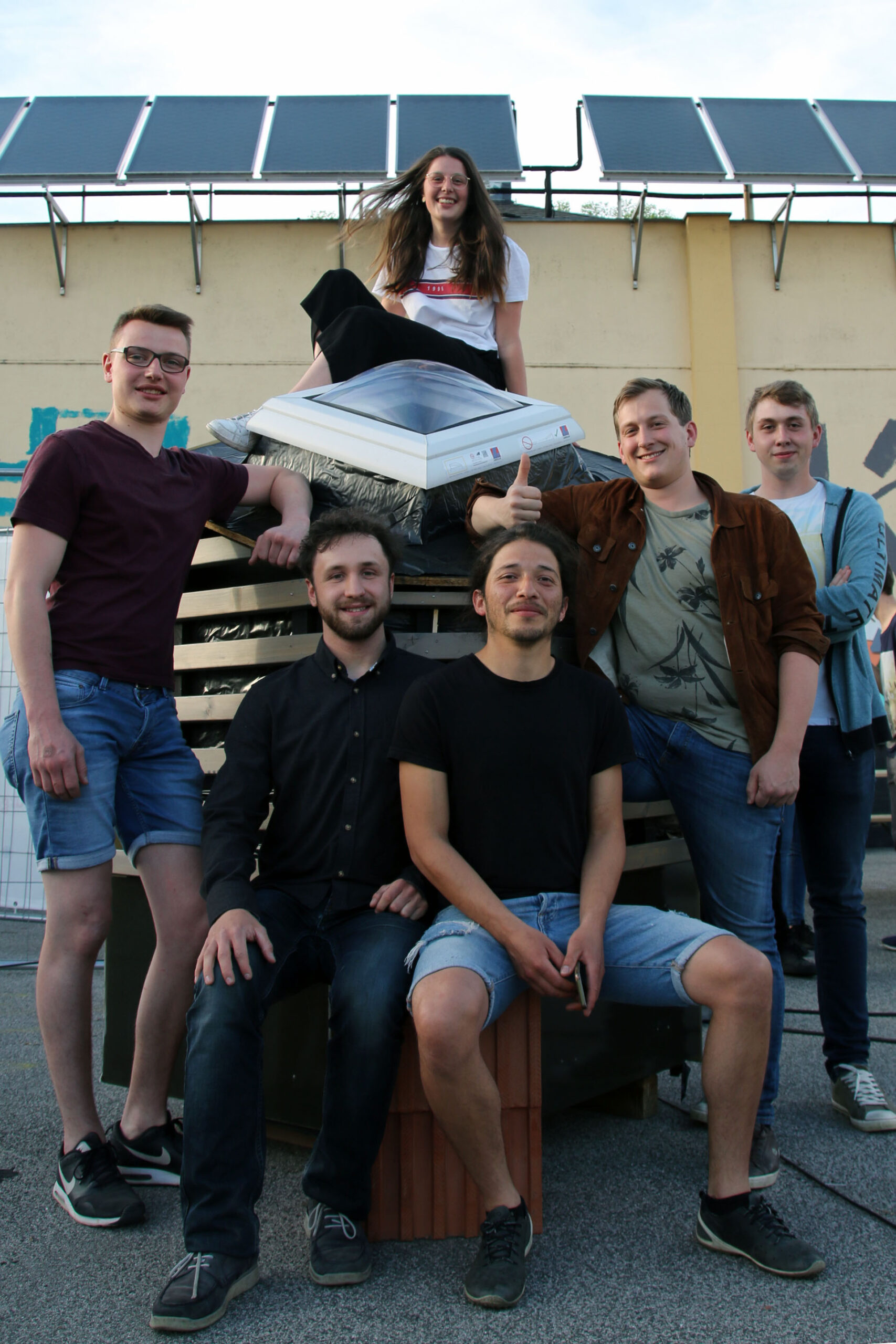 Eine Gruppe von sechs Personen, fünf Männer und eine Frau, posiert gemeinsam im Freien vor einem Solarpanel der Hochschule Coburg. Einige sitzen, andere stehen und alle lächeln in die Kamera. Sie sind lässig in Jeans und T-Shirts gekleidet und spiegeln die entspannte Atmosphäre auf dem Campus wider.