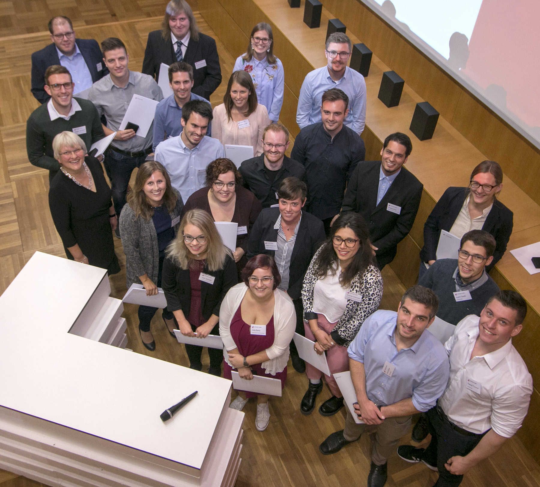 Eine Gruppe von 25 Personen in Business-Kleidung versammelt sich in einem Konferenzraum der Hochschule Coburg. Mit Zertifikaten in den Händen lächeln sie in die Kamera und fangen die freudige Atmosphäre dieser festlichen Preisverleihung ein.