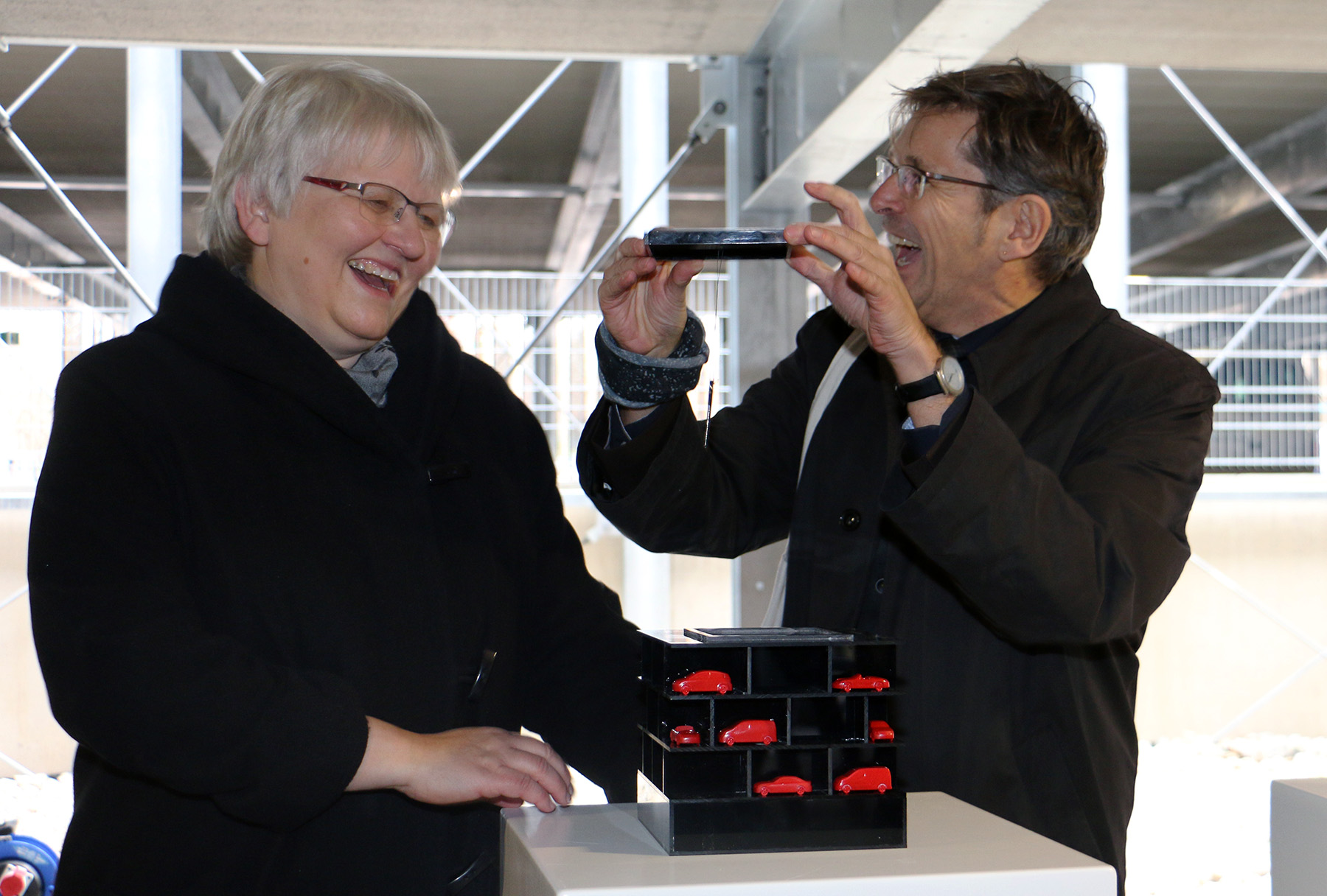 Eine Person mit kurzen weißen Haaren lächelt, während eine andere mit Brille und kurzen braunen Haaren ein Foto von einer kleinen rot-schwarzen Modellstruktur auf einem Podium macht. Im Inneren des teilweise fertiggestellten Gebäudes mit sichtbaren Metallträgern fangen sie Innovationen ein, die wahrscheinlich mit der Hochschule Coburg in Verbindung stehen.