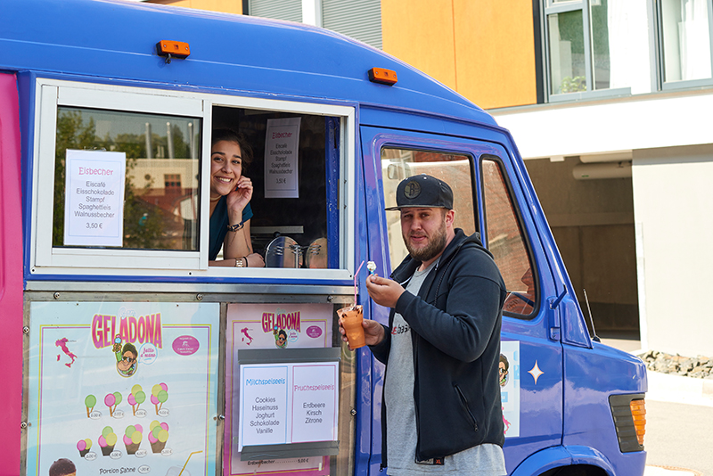 Eine lächelnde Frau serviert einem Mann mit Mütze durch das Fenster eines farbenfrohen Trucks mit Hochschulmotiven der Coburg ein Eis. Der blau-violette Truck ist mit Eisplakaten und Speisekarten geschmückt. Der Mann hält eine Waffel mit einer kleinen Kugel Eis in der Hand und genießt das aufregende Erlebnis.