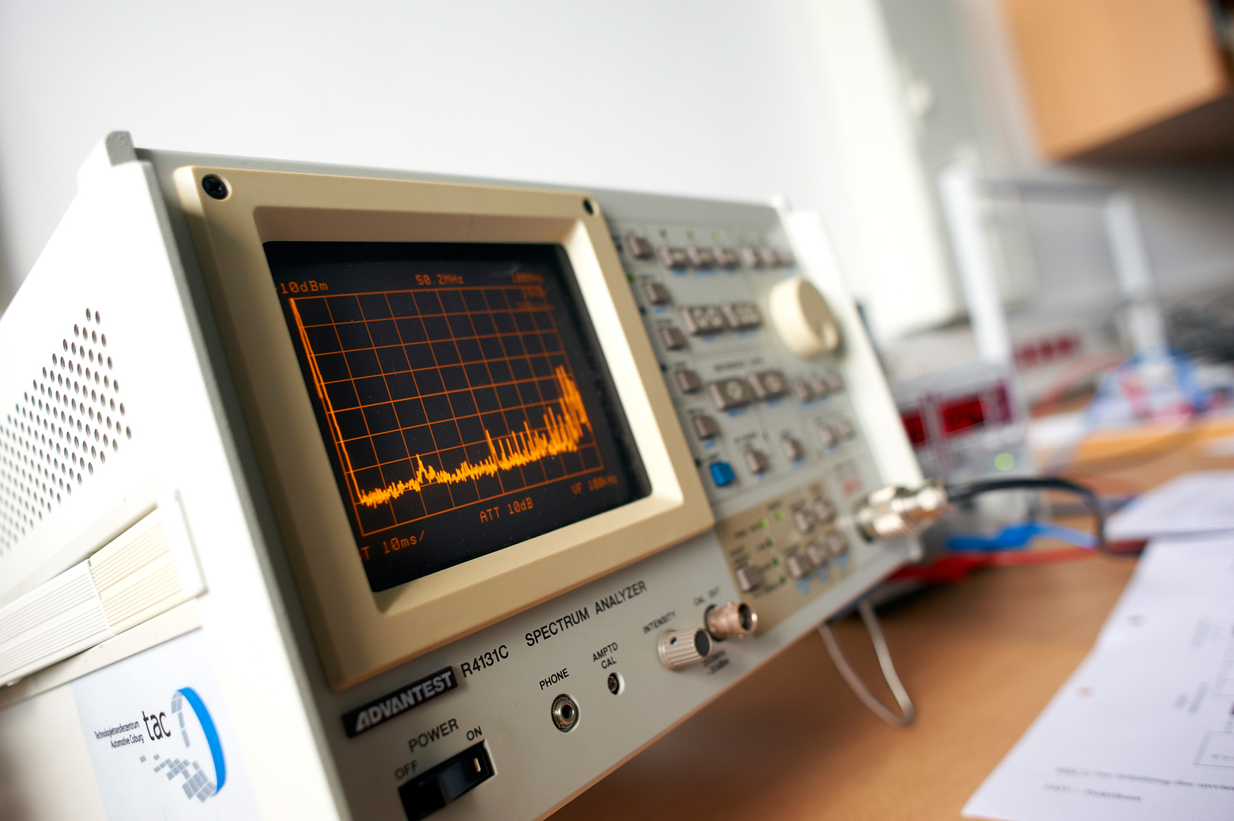 Ein Spektrumanalysator auf einem Tisch an der Hochschule Coburg zeigt auf seinem Bildschirm ein Frequenzspektrumdiagramm an. Auf dem Gerät sind zahlreiche Knöpfe und Bedienelemente zu sehen, daneben liegen verstreut Papiere.