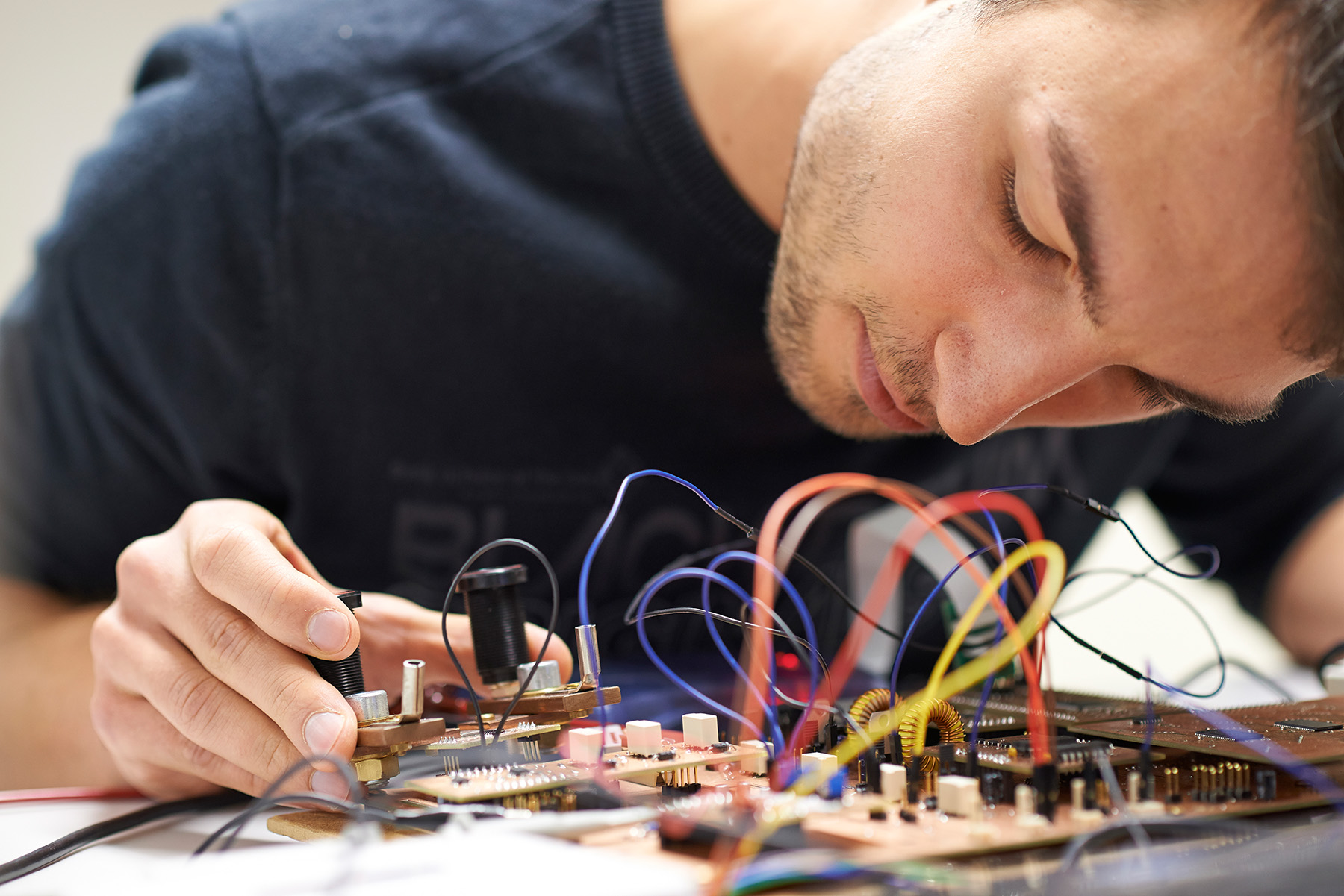 An der Hochschule Coburg konzentriert sich eine Person intensiv auf die Montage und Prüfung einer komplexen Platine mit zahlreichen bunten Drähten und Komponenten. Ihre Hände gehen behutsam mit der Elektronik um und verkörpern damit das Engagement der Universität für Präzision und Innovation im Ingenieurwesen.