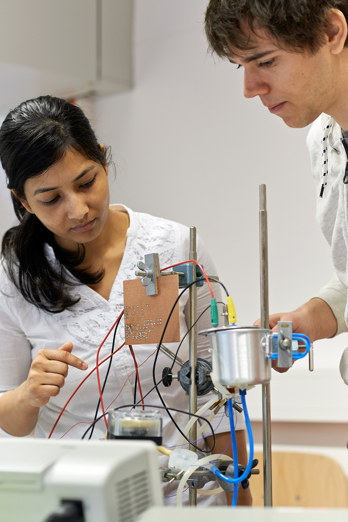 In einem Labor an der Hochschule Coburg arbeiten zwei Personen konzentriert an einem komplexen elektronischen Gerät mit Drähten und Komponenten. Einer deutet auf einen Teil des Geräts, während der andere es genau beobachtet. Beide sind konzentriert und arbeiten zusammen, was ihr Engagement für Innovation zeigt.