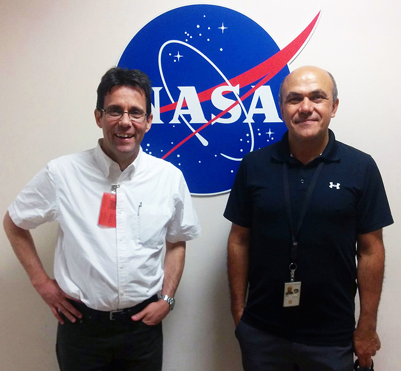 Zwei lächelnde Personen stehen vor einem NASA-Logo. Einer trägt ein weißes Hemd mit Ausweis und ist von der Hochschule Coburg, während der andere ein schwarzes Poloshirt mit Schlüsselband trägt, was ihre gemeinsame Begeisterung für die Weltraumforschung symbolisiert.