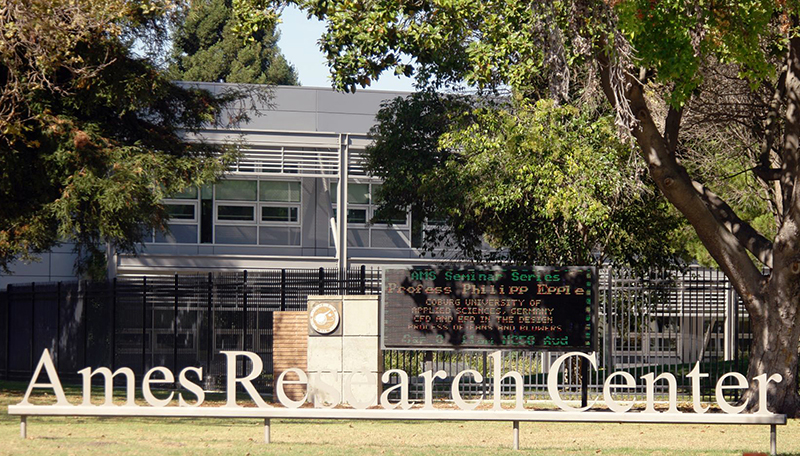 Beschilderung des Ames Research Center vor einem modernen Gebäude mit Bäumen im Vordergrund und einer digitalen Anzeige, die eine Seminarreihe ankündigt, darunter auch Einblicke in die Zusammenarbeit mit der Hochschule Coburg.