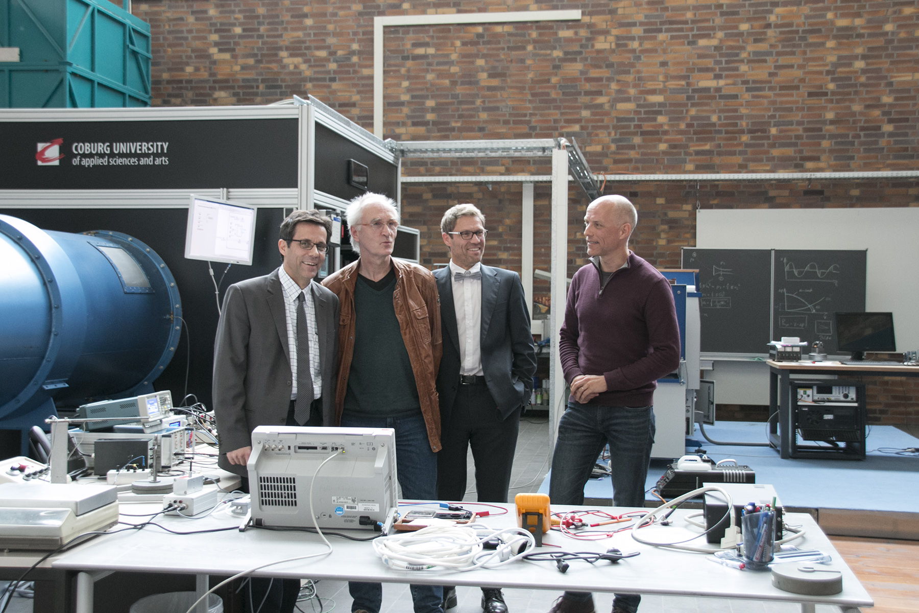 Vier Männer stehen in einer Werkstatt der Hochschule Coburg, umgeben von elektronischen Geräten und Maschinen. Sie scheinen in ein Gespräch vertieft zu sein. Eine blaue Maschine und eine Tafel voller Diagramme bestimmen die Szenerie.