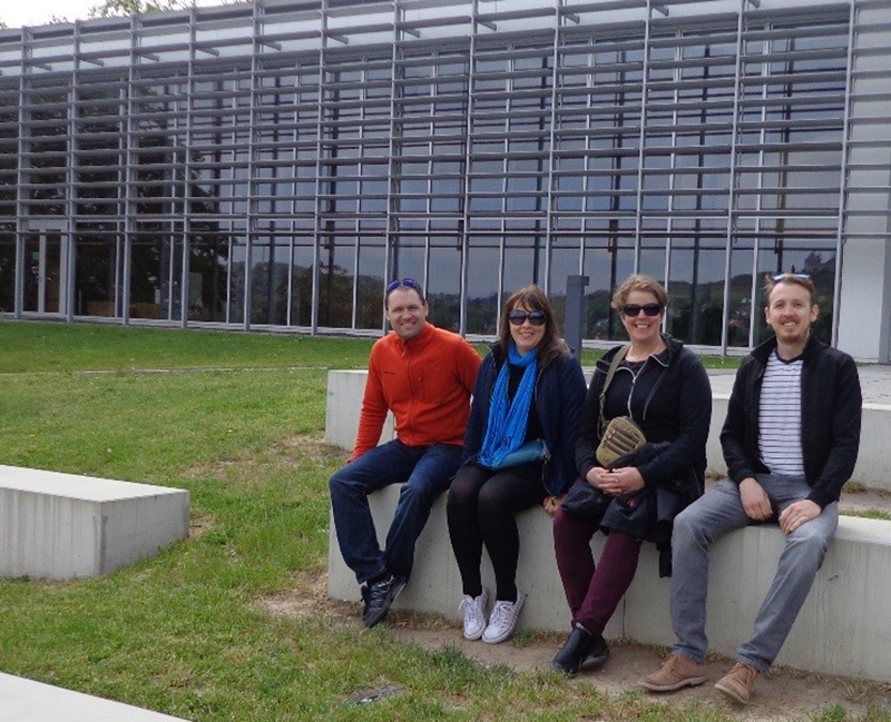 Auf einer Grünfläche vor der modern gestalteten Hochschule Coburg mit ihren großen Glasfenstern und horizontalen Metallgittern sitzen vier Menschen auf Betonbänken. Sie wirken entspannt und sind leger gekleidet.