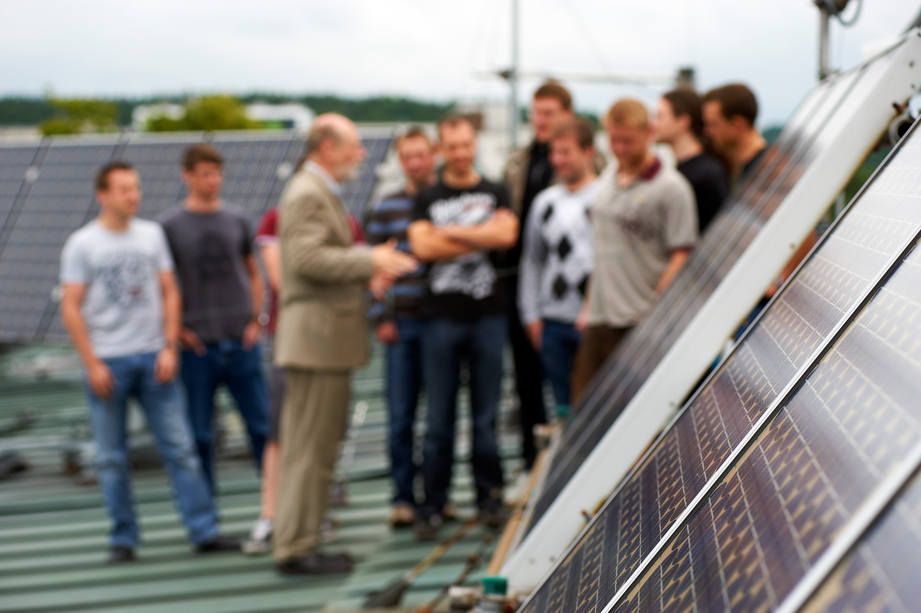 Eine Gruppe von Menschen steht auf einem Dach der Hochschule Coburg und hat sich um einen Mann im Anzug versammelt, der auf eine Reihe von Solarmodulen zeigt. Die Solarmodule sind deutlich im Vordergrund zu sehen und ragen in den Himmel.