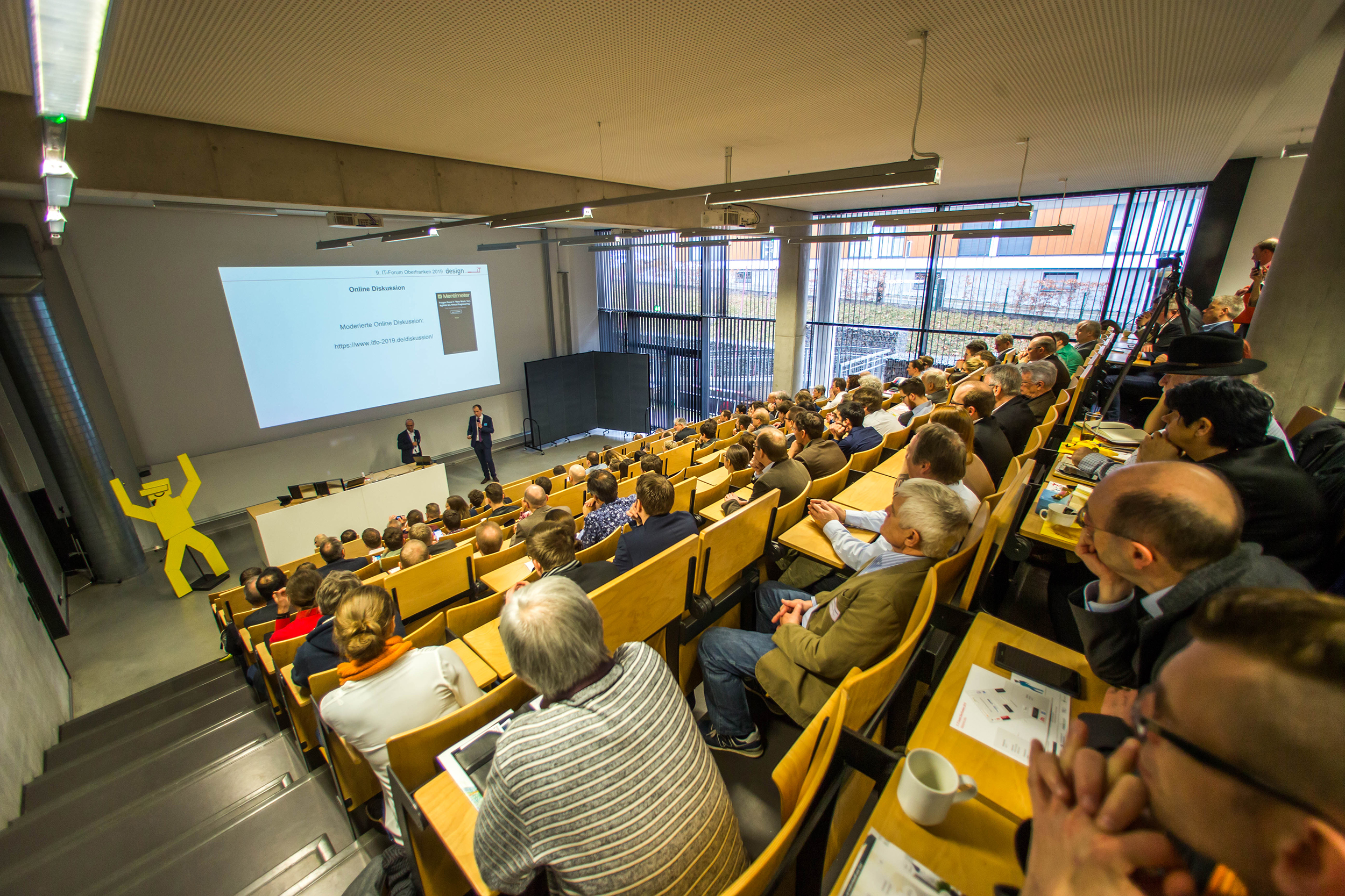 In einem vollen Hörsaal der Hochschule Coburg sitzen die Teilnehmer in Reihen aus Holzstühlen. Zwei Vortragende stehen vorne neben einer großen Leinwand, auf der eine Präsentation läuft. Links von ihnen ist eine gelbe Statue zu sehen.