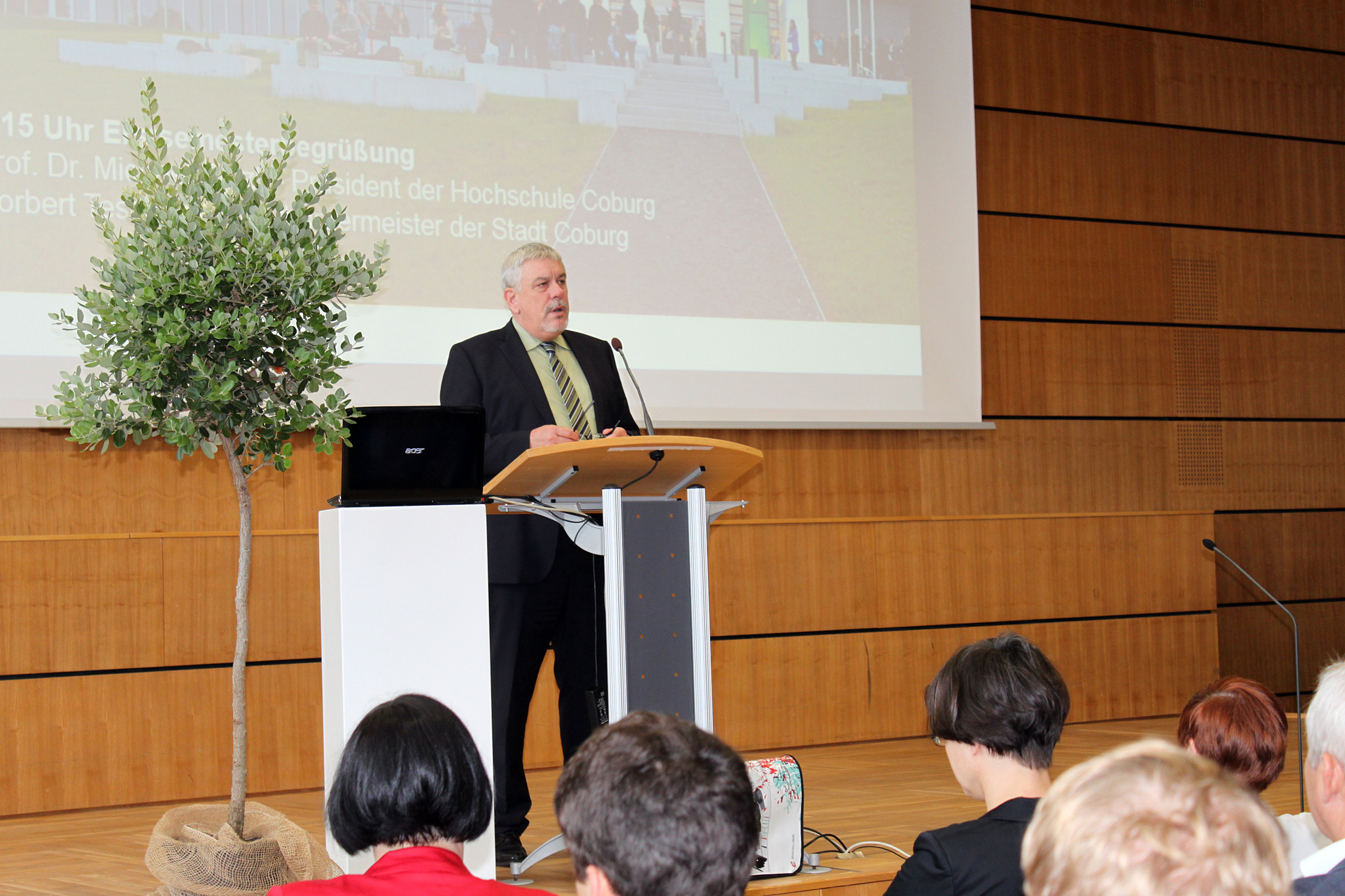 Ein Mann spricht an einem Podium in einem Konferenzraum mit Holzwänden der Hochschule Coburg. Neben ihm steht ein kleiner Baum, das Publikum sitzt aufmerksam da. Im Hintergrund läuft auf einer Projektionsfläche eine teilweise sichtbare Diashow.