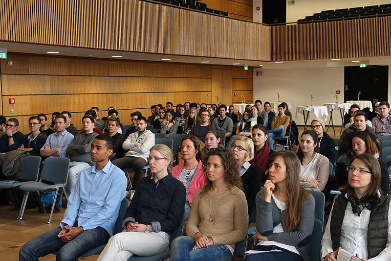 Eine bunt gemischte Gruppe sitzt in einem Hörsaal der Hochschule Coburg und hört aufmerksam einer Präsentation zu. Der Raum hat Holzwände und eine hohe Decke. Auf einem Zwischengeschoss sind weitere Sitzplätze zu sehen.