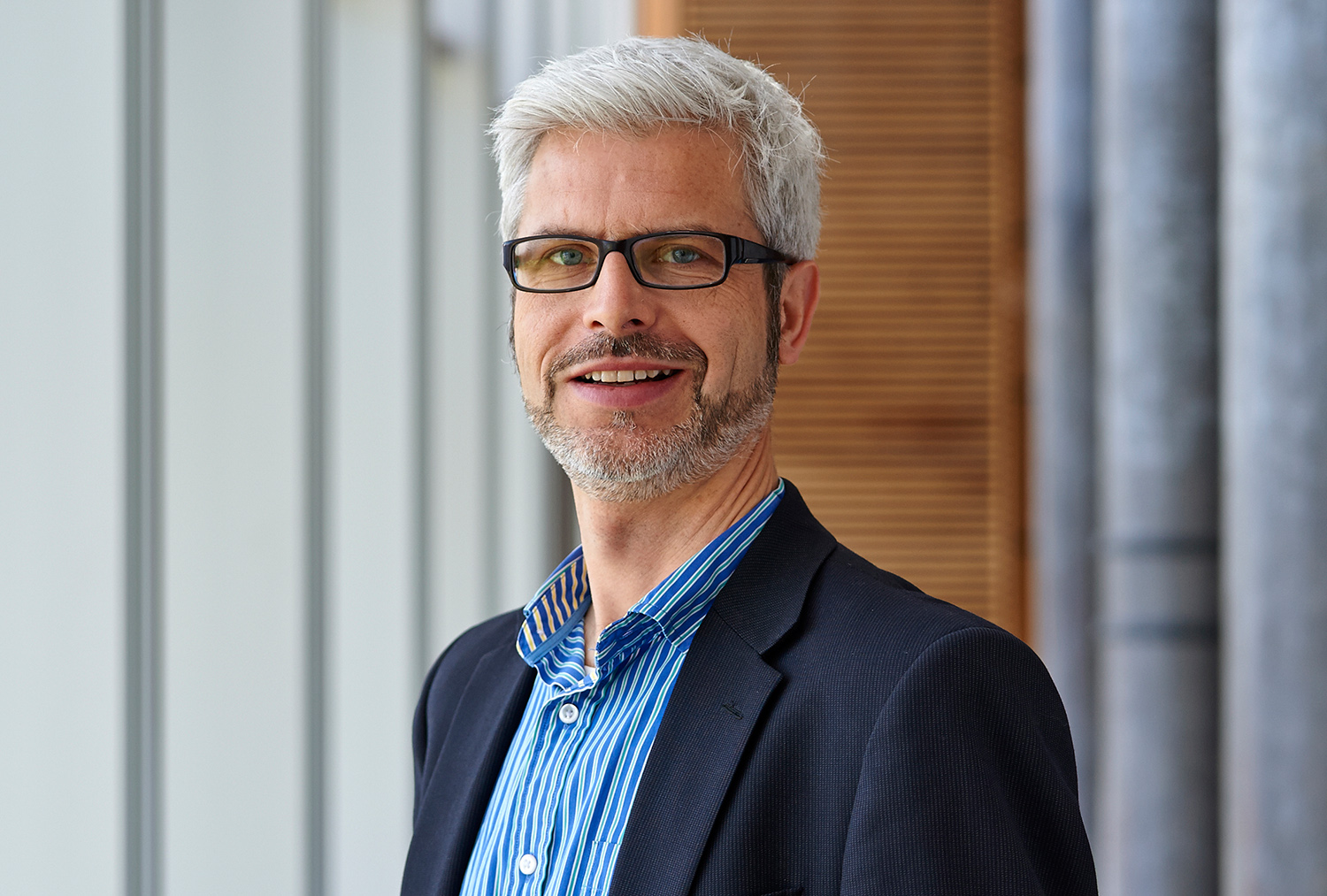 Ein Mann mit kurzen grauen Haaren und grauem Bart, einer Brille und einem dunklen Blazer über einem blau gestreiften Hemd steht drinnen neben einem Fenster. Er lächelt in die Kamera und verkörpert damit den professionellen Geist der Hochschule Coburg.