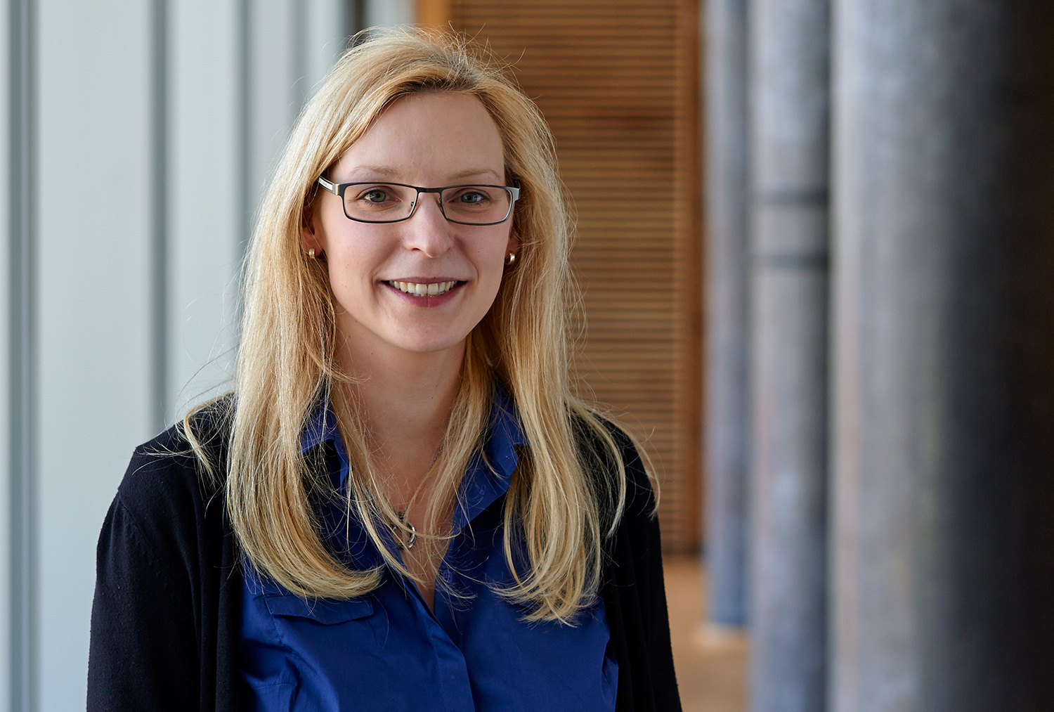 Eine Frau mit langen blonden Haaren und Brille lächelt, trägt ein blaues Hemd und einen schwarzen Pullover. Sie steht in einem Flur, im Hintergrund ist eine Holzvertäfelung zu sehen, die an die elegante Architektur der Hochschule Coburg erinnert.